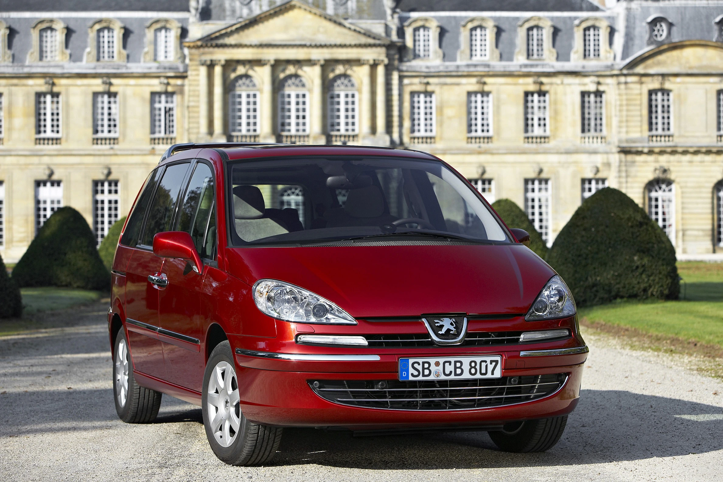 Roter Peugeot Minivan vor einem historischen Gebäude mit vielen Fenstern und einem symmetrischen Garten im Hintergrund.