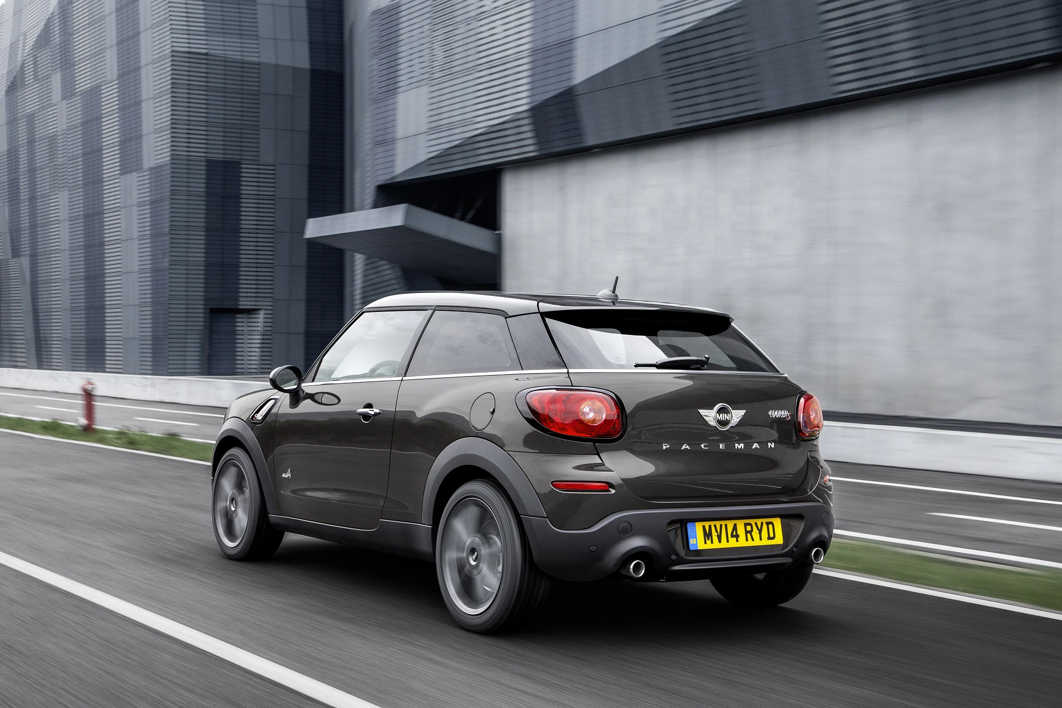 Ein grauer Mini Paceman fährt auf einer Straße neben einem modernen Gebäude. Lizenznummer auf gelbem Schild sichtbar.