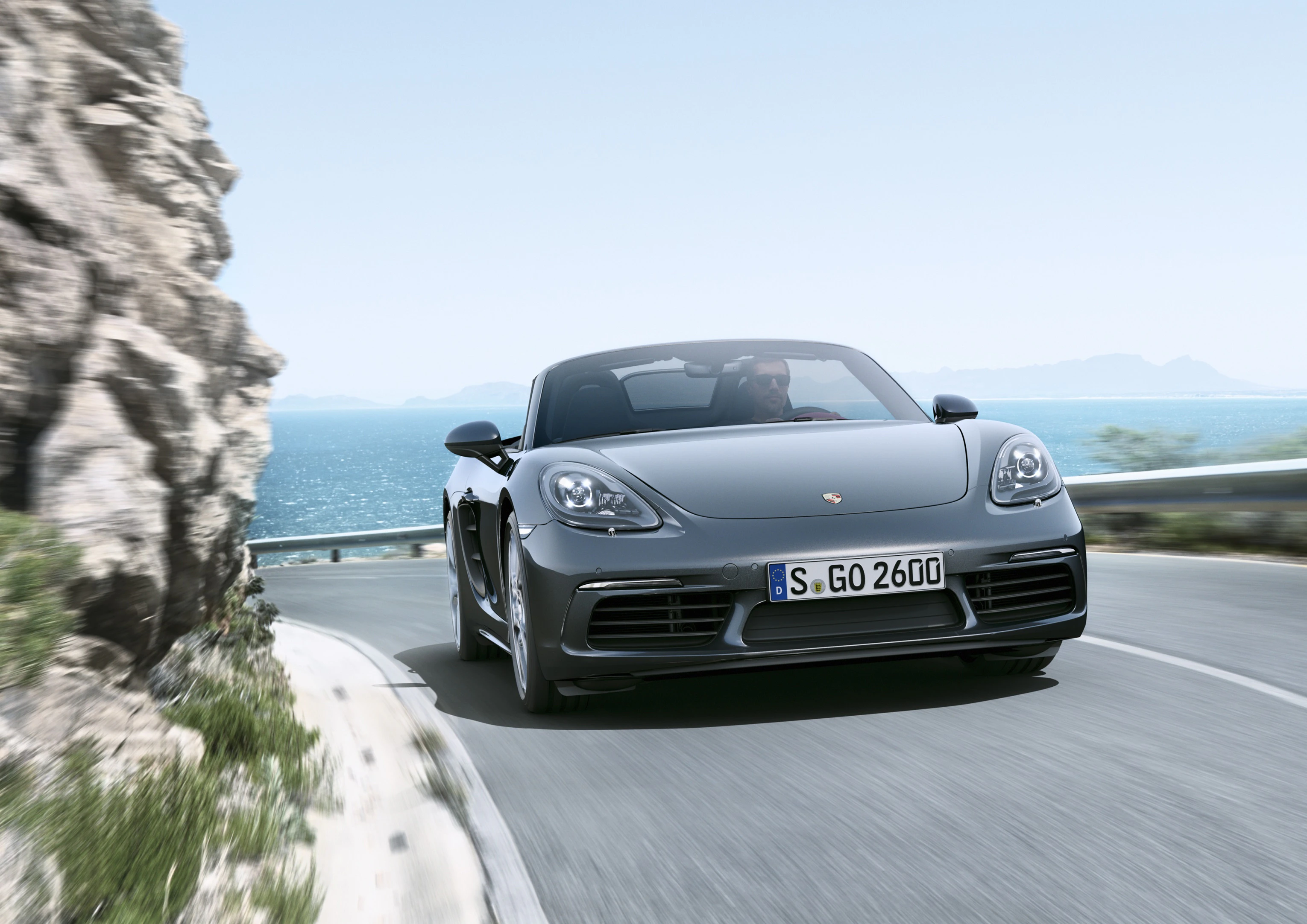 Ein grauer Cabrio-Sportwagen fährt an einer kurvigen Küstenstraße entlang, im Hintergrund sind Meer und Felsen zu sehen.