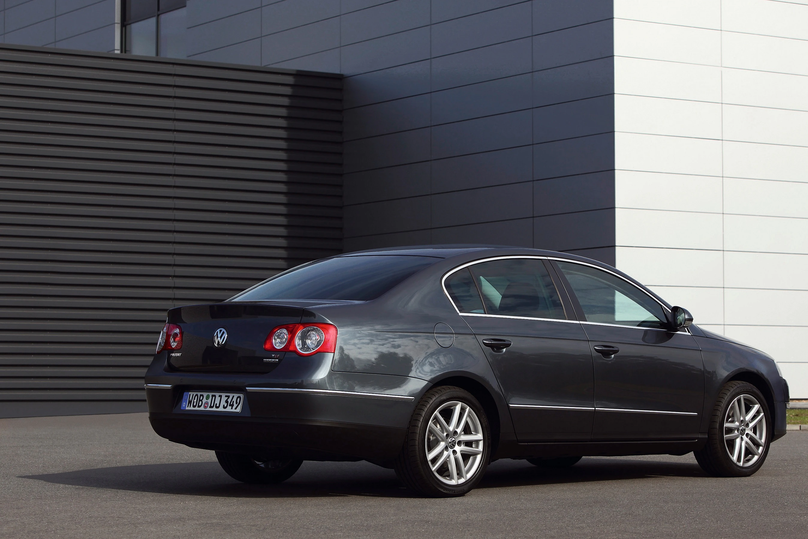 Schwarzer Volkswagen Passat, Heckansicht, auf einem Parkplatz vor einem modernen Gebäude mit grauen und weißen Paneelen.