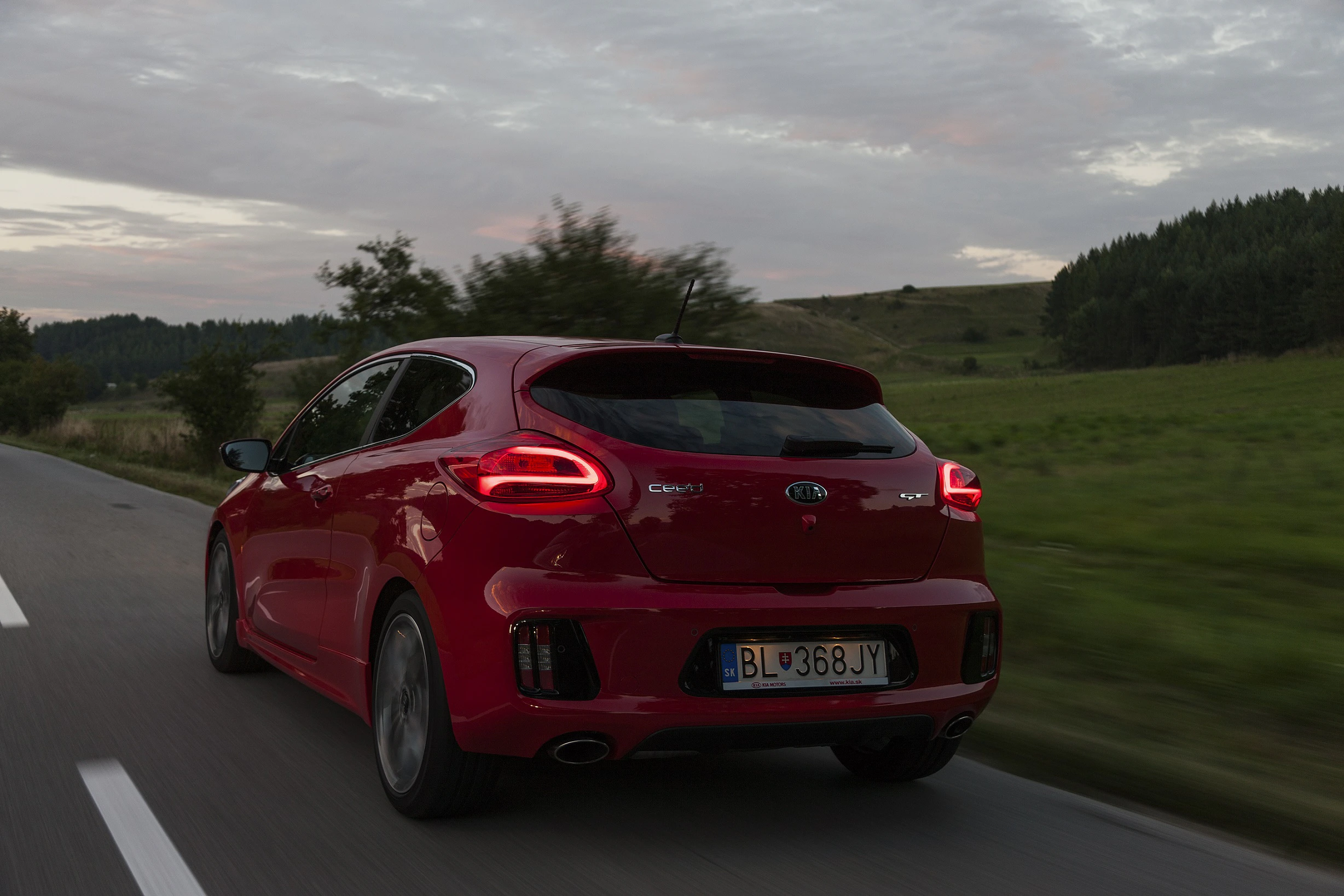 Roter Kia Ceed GT fährt bei Sonnenuntergang auf einer Landstraße durch grüne, ländliche Umgebung. Rücklichter leuchten.