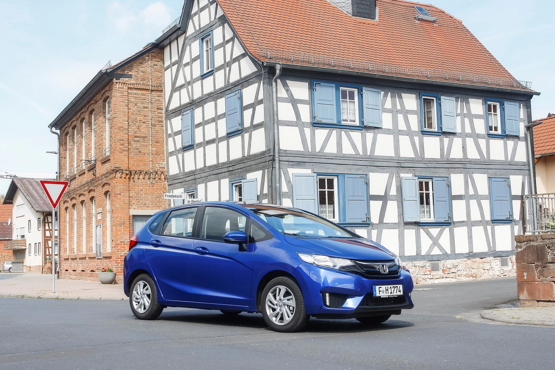 Blaues Auto parkt auf einer Straße vor einem Fachwerkhaus und einem Backsteinhaus bei sonnigem Wetter.