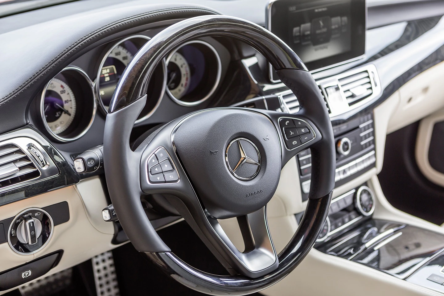 Lenkrad eines Mercedes-Benz mit silbernem Logo, umgeben von einem modernen Armaturenbrett mit eingebautem Display und mehreren Bedienungselementen.