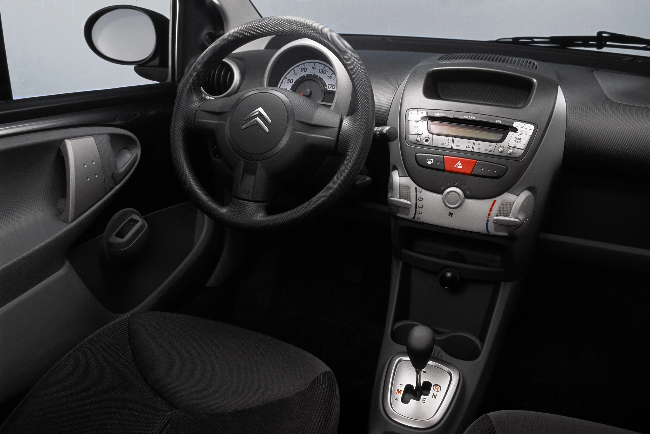 Innenraum eines Autos mit Lenkrad links, Armaturenbrett, Schalthebel und verschiedenen Bedienelementen; modernes, schlichtes Design.