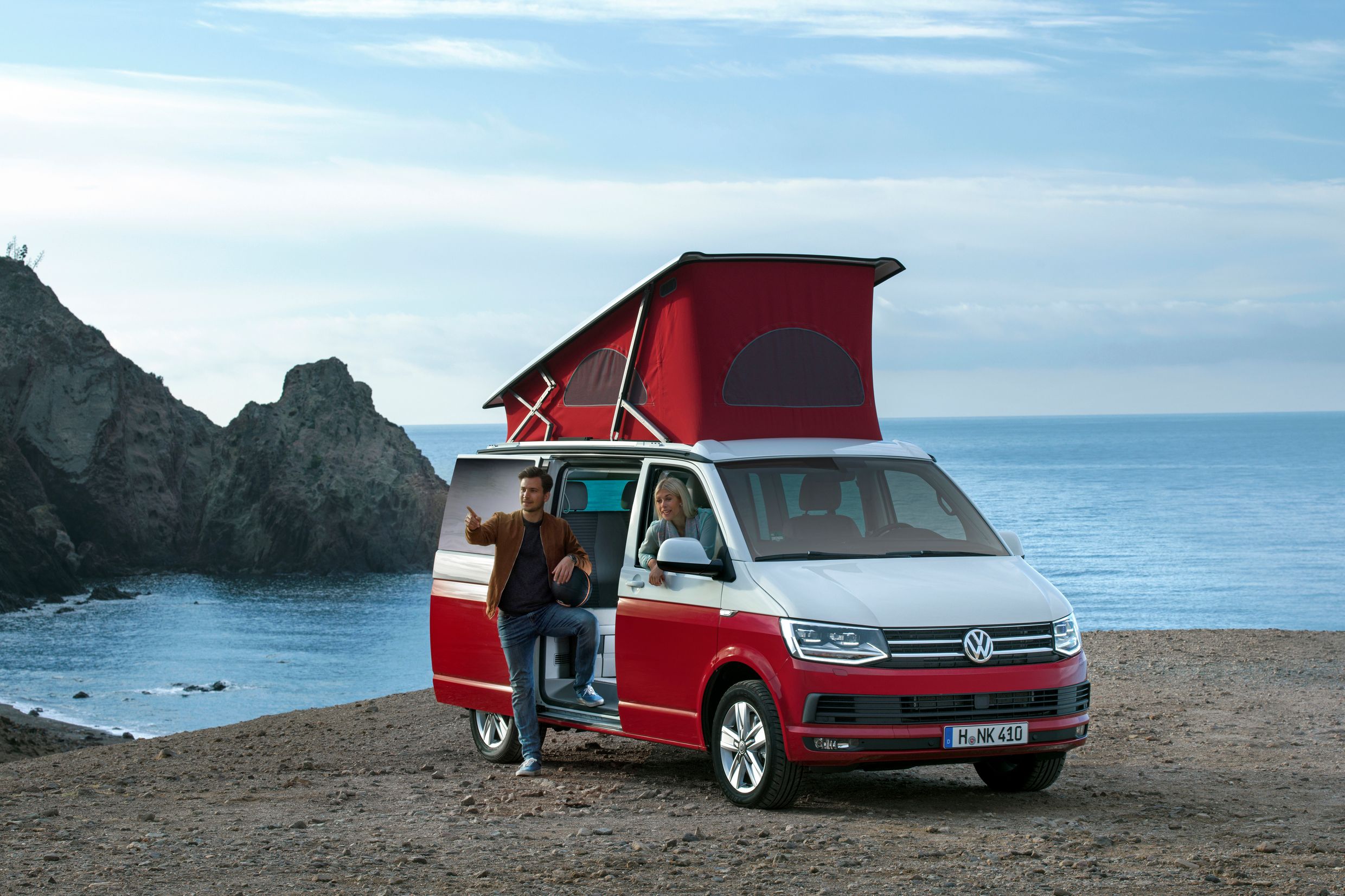 Roter VW-Campingbus mit ausgefahrenem Dach am Strand. Zwei Menschen sitzen im Auto mit Blick auf das Meer und die Felsen im Hintergrund.