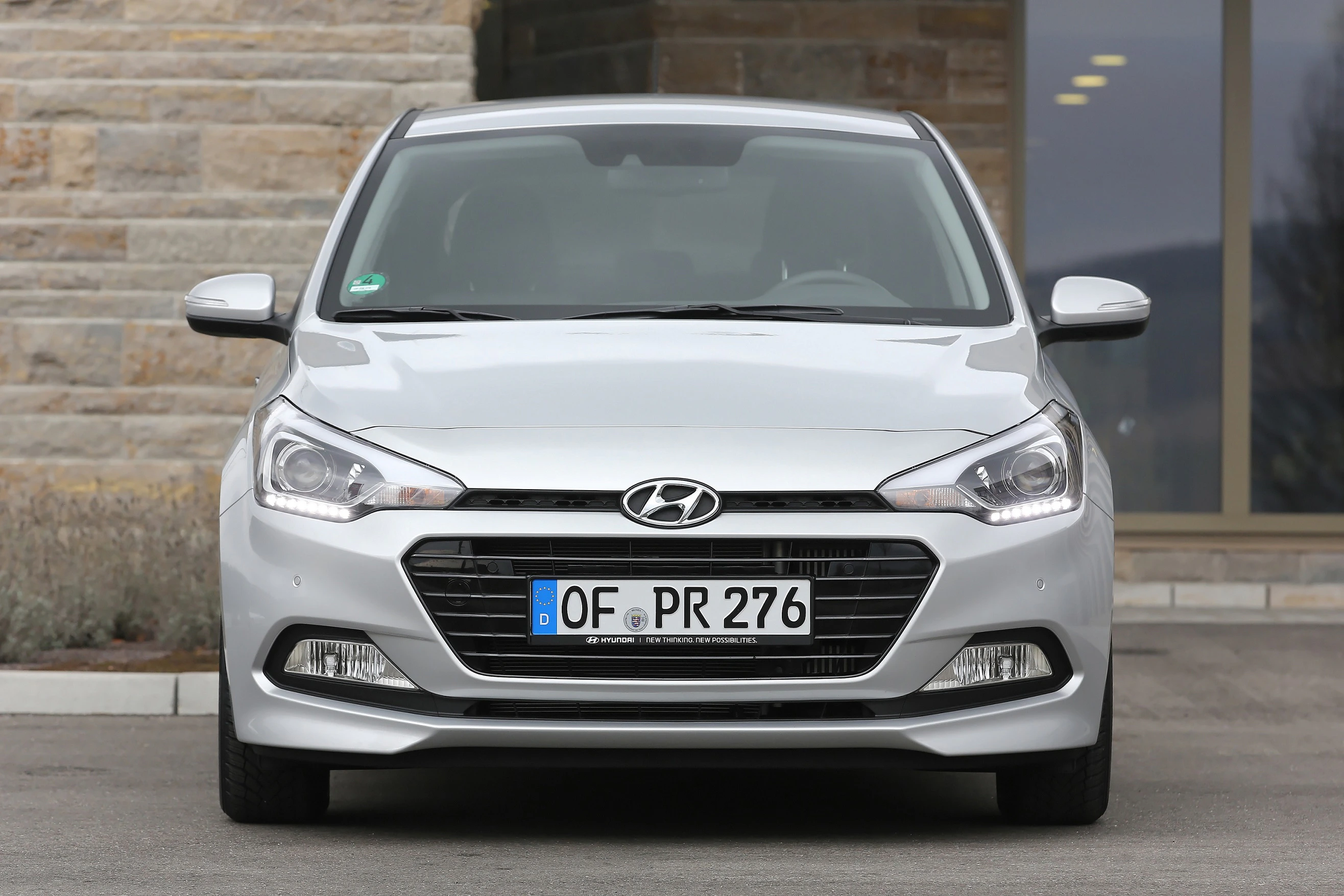 Silberner Hyundai i20, Vorderansicht, parkend vor einem Gebäude mit Glasfenstern und Steinwänden.