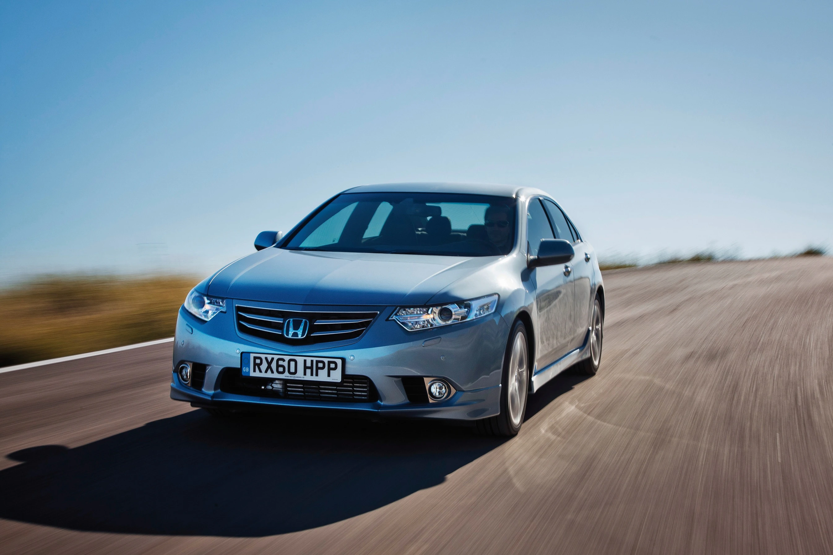 Silberner Honda-Acura fährt schnell auf leerer, kurviger Straße unter blauem Himmel.