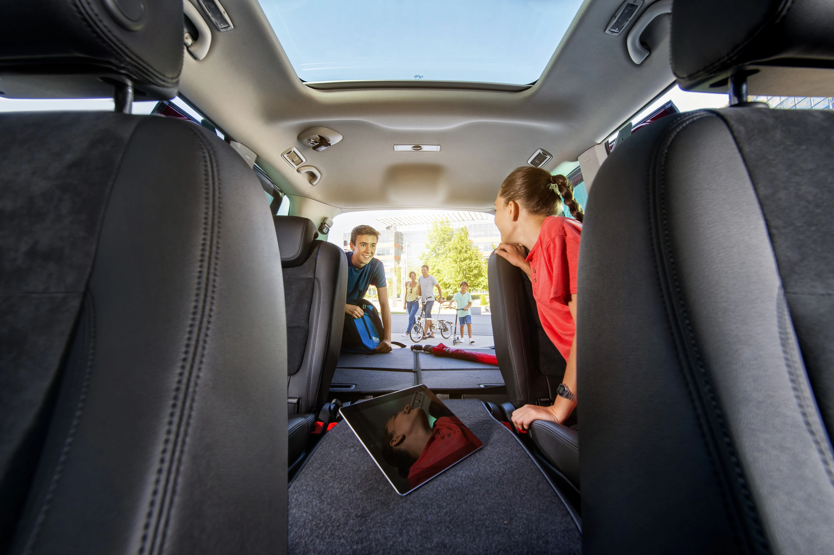Blick von innen auf den Kofferraum eines Autos mit zwei Kindern, die hineinsehen. Ein Tablet liegt auf dem umgeklappten Sitz.