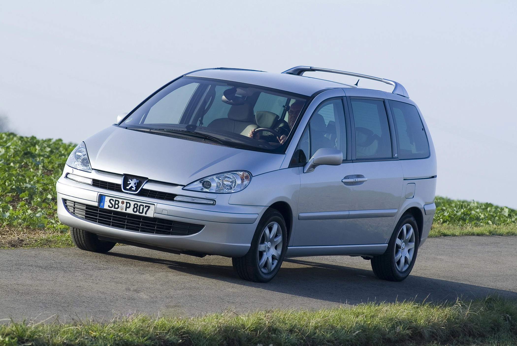 Silberner Peugeot Minivan fährt auf einer Landstraße mit grünen Feldern im Hintergrund. Fahrer sitzt am Steuer, sonniger Tag.