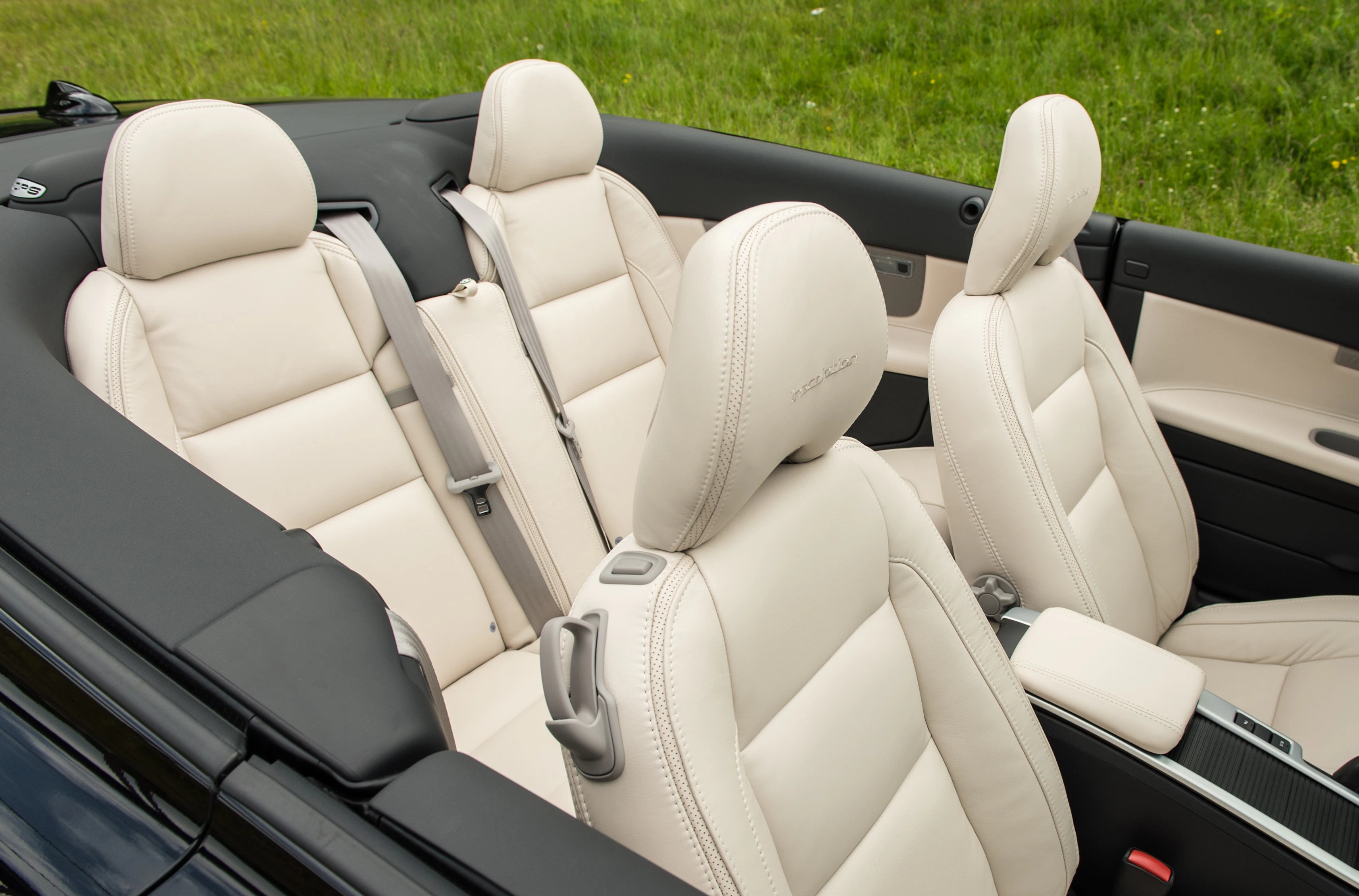 Innenraum eines Cabriolets mit beigen Ledersitzen und Armlehnen, umgeben von grünem Gras im Hintergrund.
