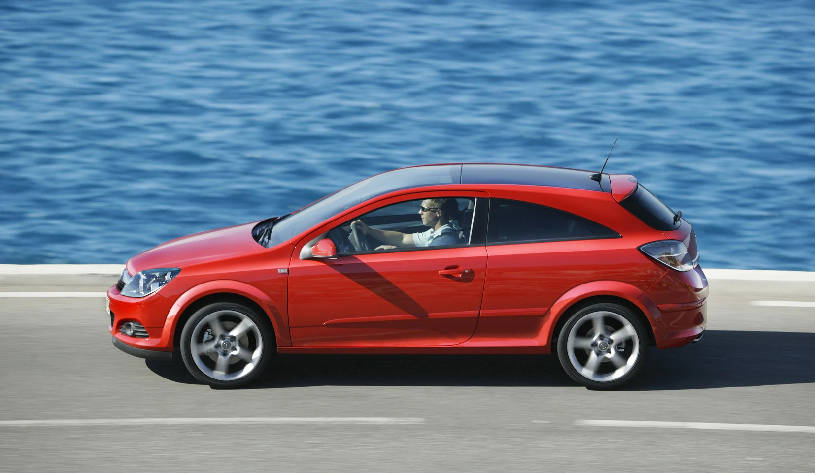 Rotes Auto fährt auf einer Küstenstraße entlang, im Hintergrund das blaue Meer. Fahrer am Steuer, Sommerwetter.
