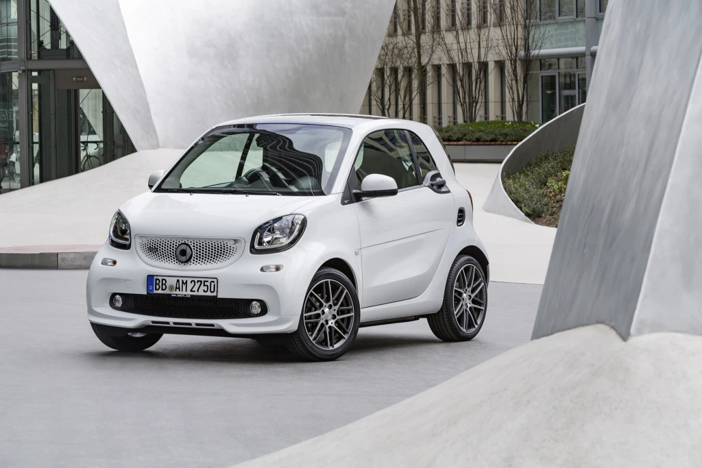 Kleiner weißer Smart Fortwo parkt in modernem, urbanem Umfeld mit futuristischen Gebäudeelementen im Hintergrund.