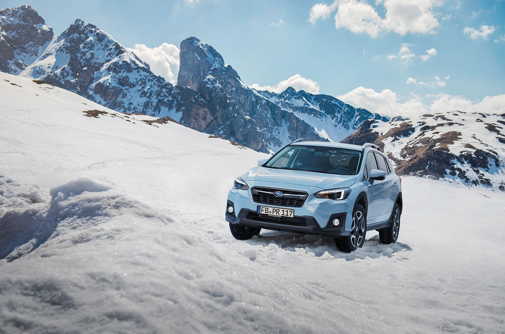 Ein silberner SUV fährt durch eine verschneite Berglandschaft bei klarem Himmel. Majestätische Gipfel im Hintergrund.
