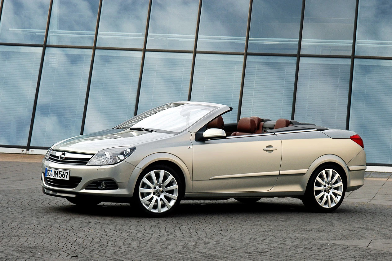 Silbernes Cabrio vor moderner Glasfassade, seitlich aufgenommen. Verdeck offen, zeigt braune Ledersitze, elegantes Design.