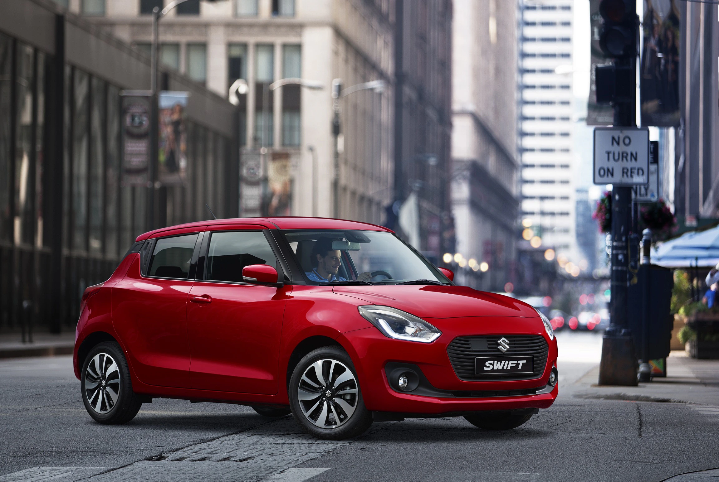 Roter Suzuki Swift steht auf einer urbanen Straße mit Hochhäusern im Hintergrund und einem Verkehrsschild "No Turn On Red".