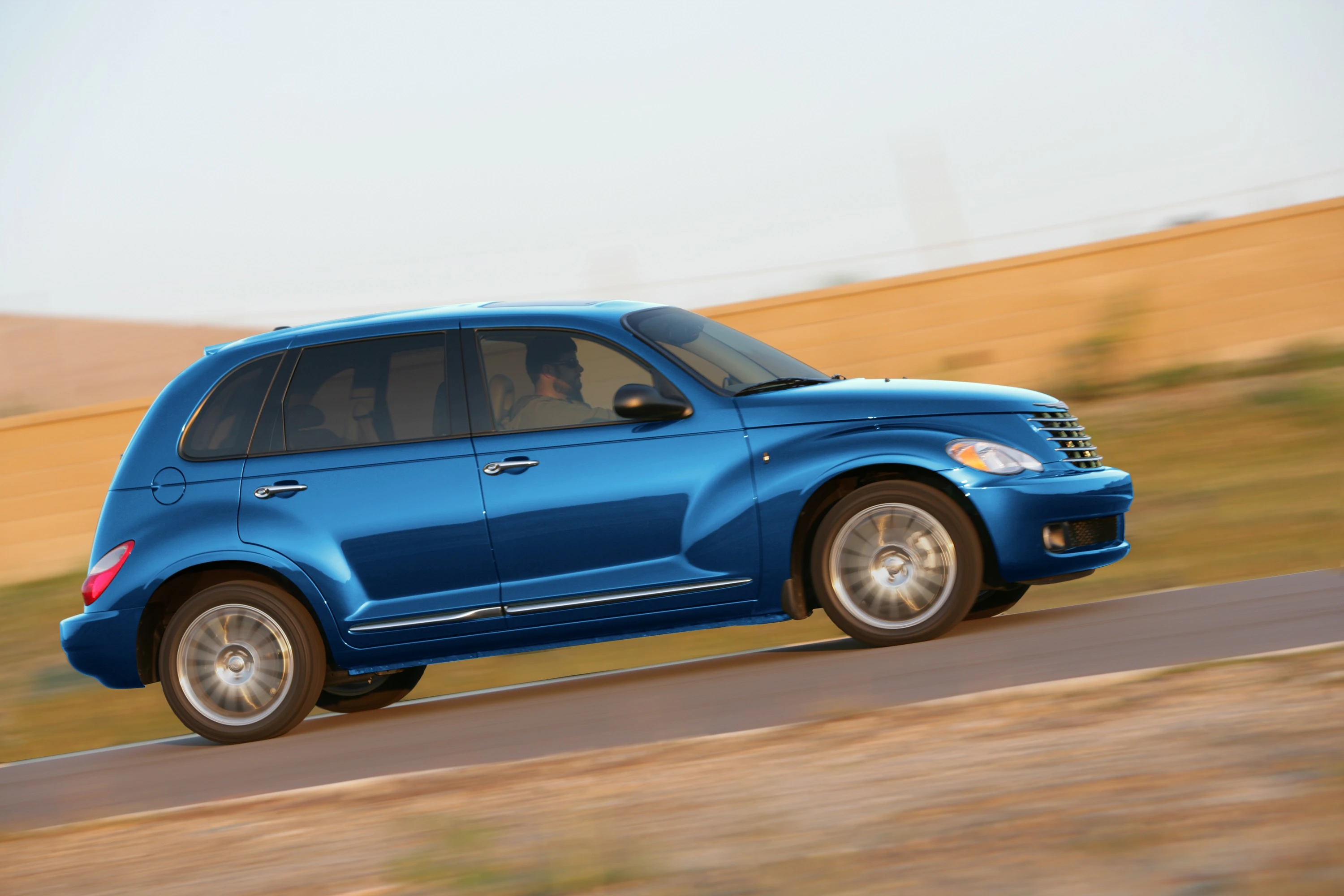 Blauer PT Cruiser fährt auf einer Straße durch eine sandige Landschaft bei Tageslicht, mit zwei Personen im Auto.