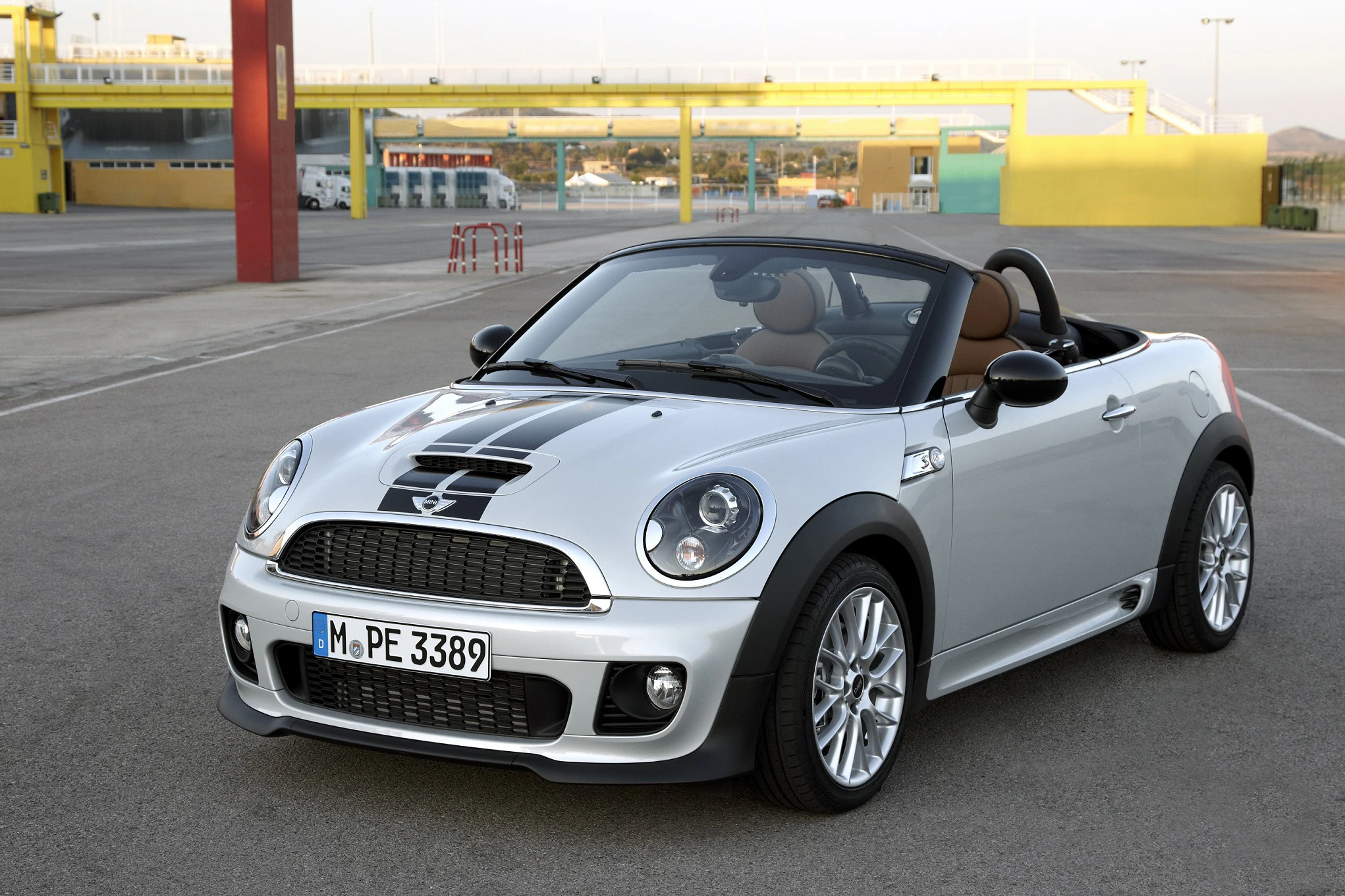 Silberner Mini Cooper Cabrio mit schwarzen Streifen, geparkt auf offenem Asphalt. Hintergrund mit bunter Architektur.