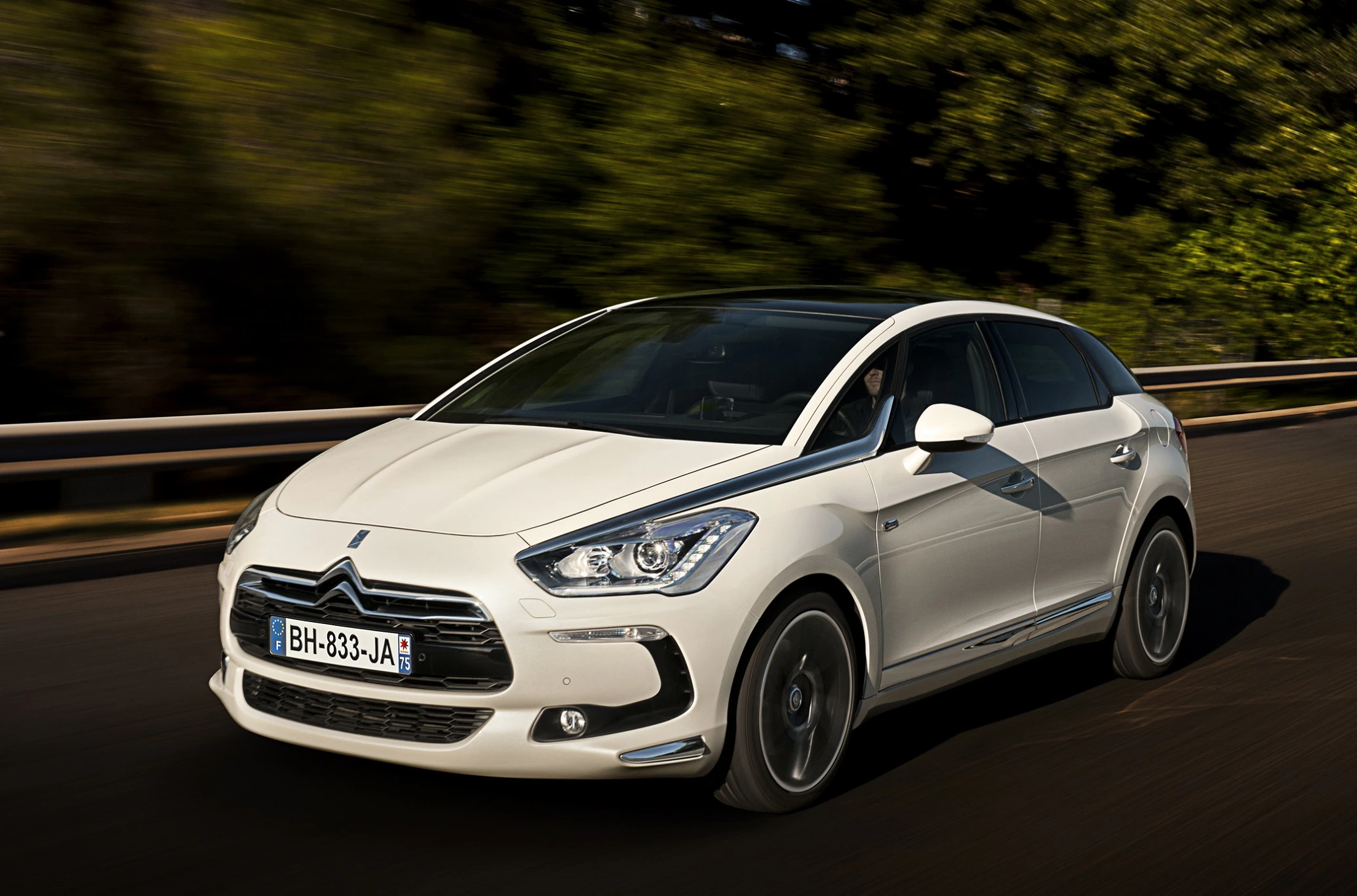 Weißer Citroën DS5 fährt auf einer Straße mit grünen Bäumen im Hintergrund.