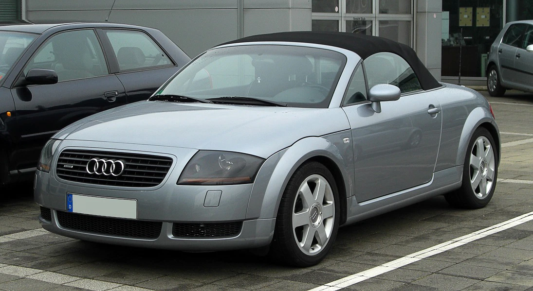 Silberner Audi TT Cabrio parkt auf einem Parkplatz vor einem Gebäude. Auto mit schwarzen Reifen und geschlossenem Verdeck.