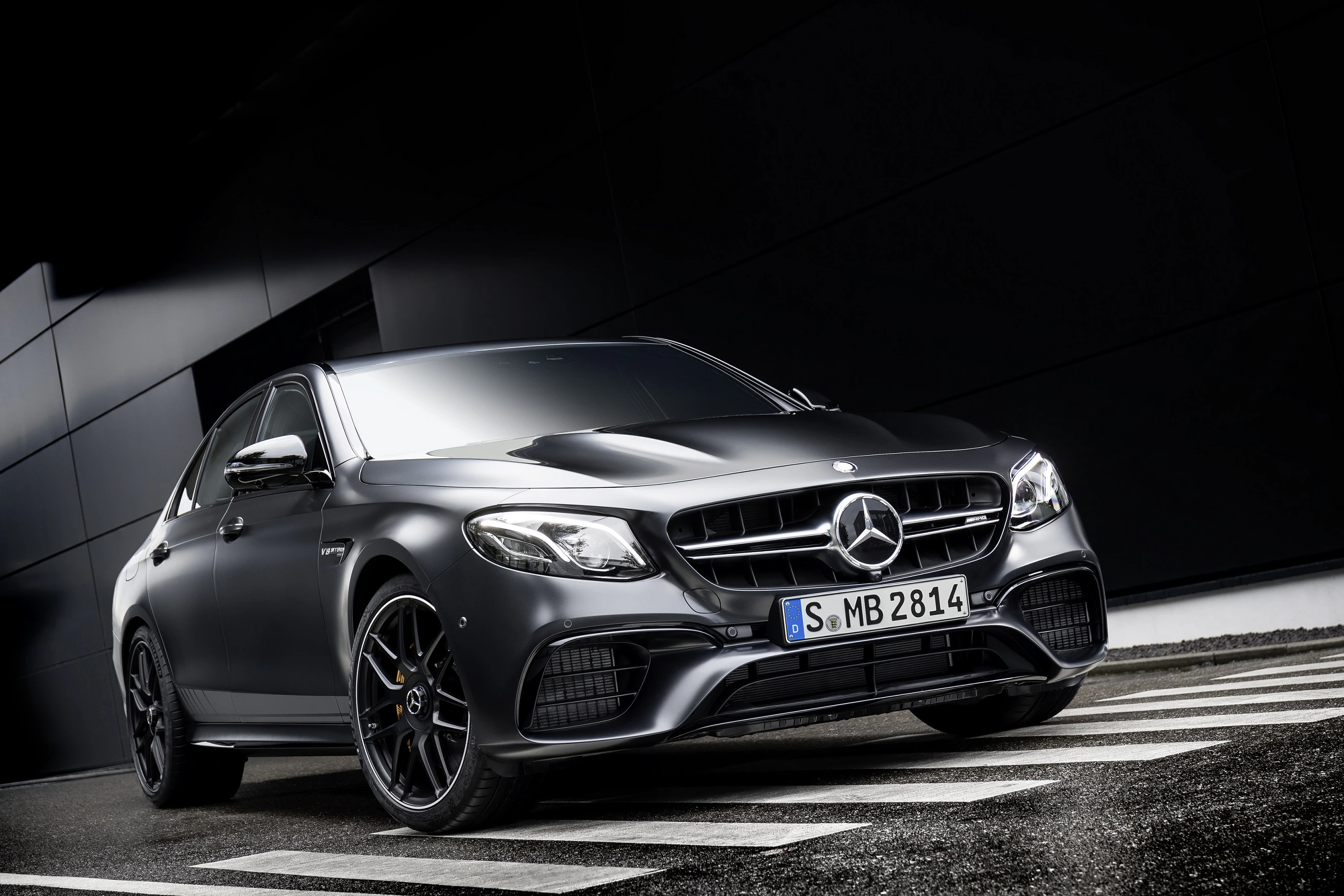 Mattschwarzer Mercedes-Benz auf nasser Straße, seitliche Frontansicht, Gebäude im Hintergrund.