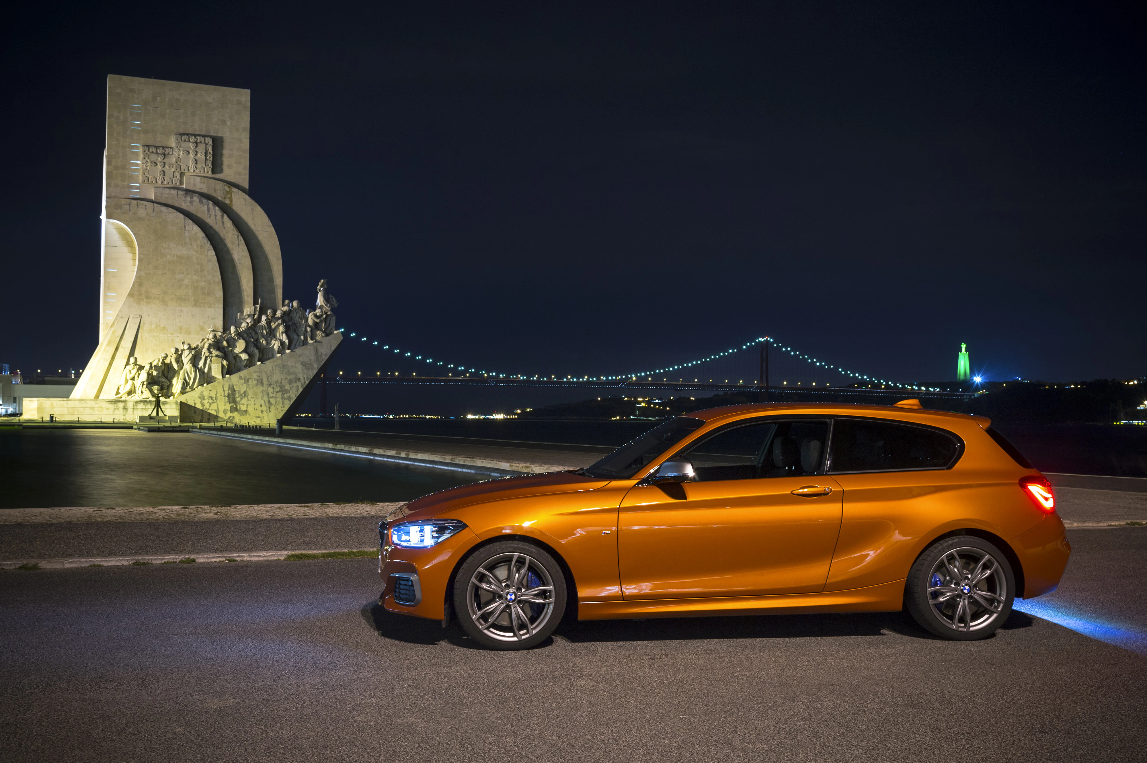 Ein orangefarbenes Auto vor dem Denkmal der Entdeckungen in Lissabon, mit Brücke und beleuchteter Stadt im Hintergrund bei Nacht.