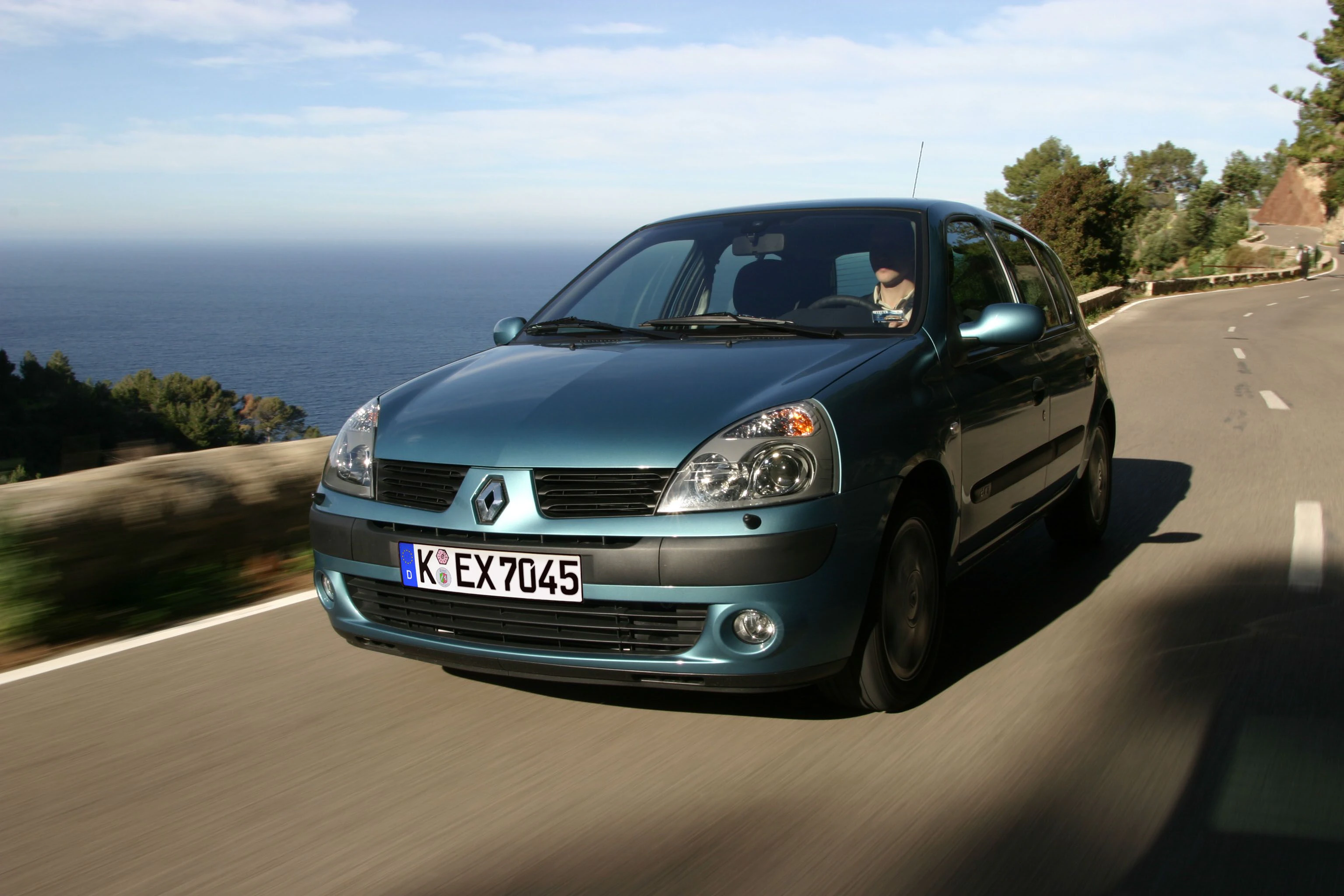 Ein blauer Renault Clio fährt auf einer kurvigen Straße entlang der Küste, mit Meer und Bäumen im Hintergrund unter klarem Himmel.