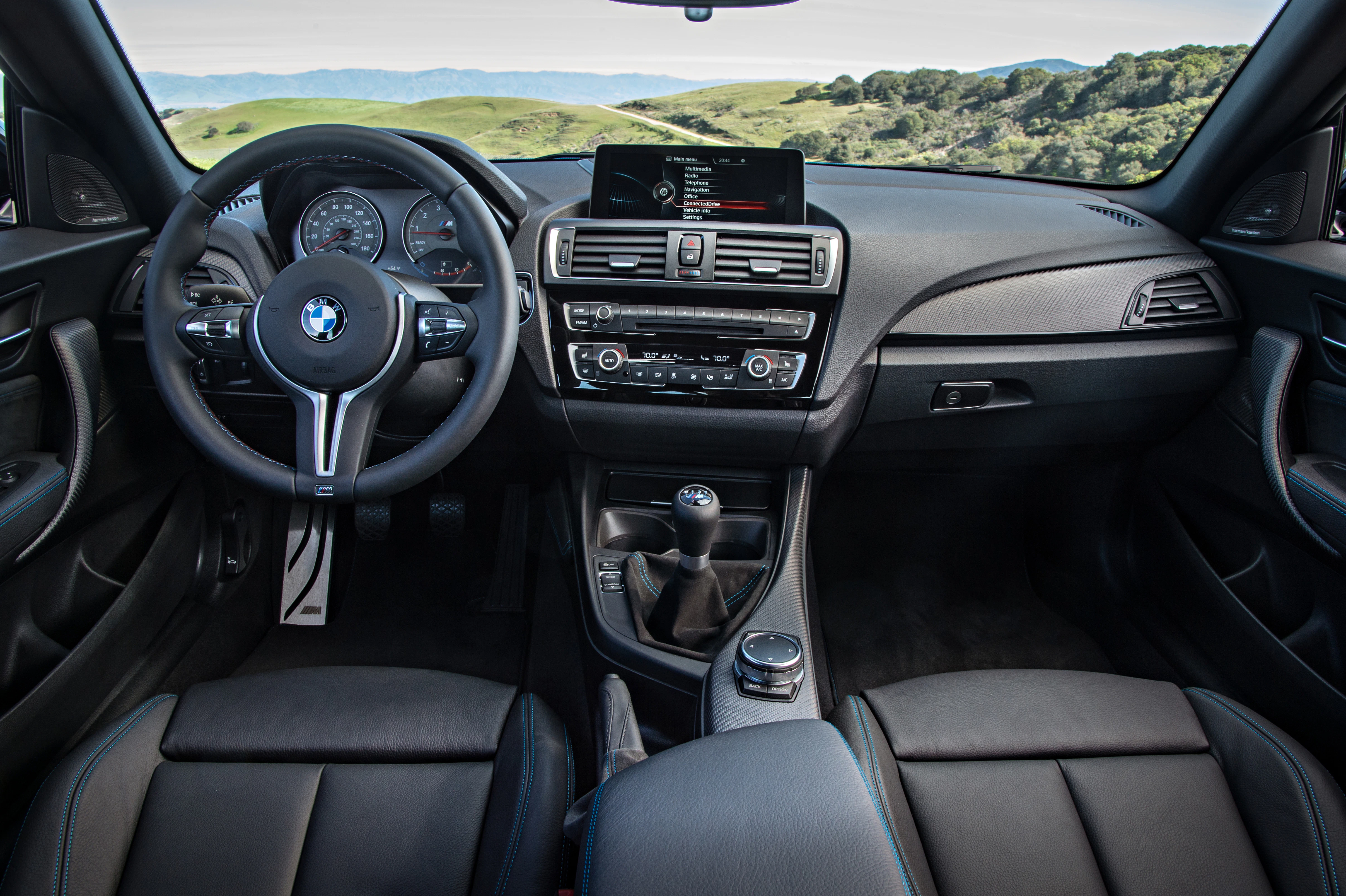 Innenraum eines BMW mit Lederpolstern, Lenkrad, manueller Gangschaltung und Display. Blick durch die Windschutzscheibe auf grüne Hügel.
