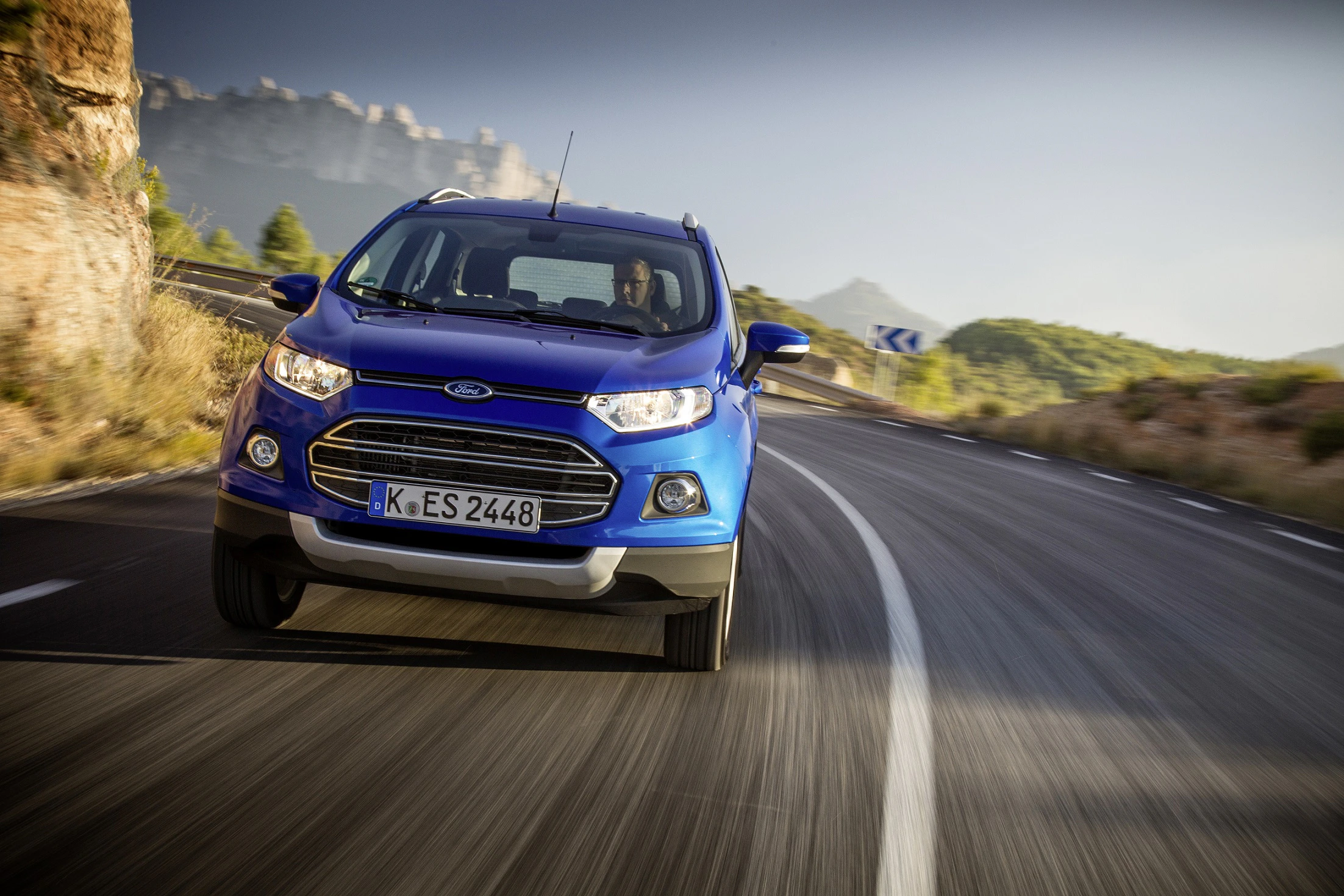 Blauer SUV fährt auf kurviger Landstraße in bergiger Landschaft bei sonnigem Wetter. Fahrzeug ist von vorn in Bewegung zu sehen.