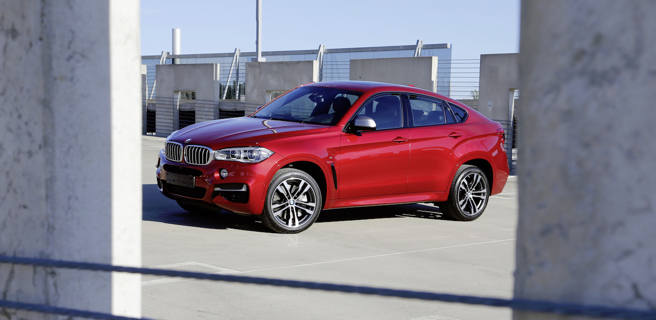 Roter BMW-SUV auf einem Parkplatz, umgeben von Betonpfeilern, bei klarem Himmel. Luxuriöses Design, sportliche Felgen.