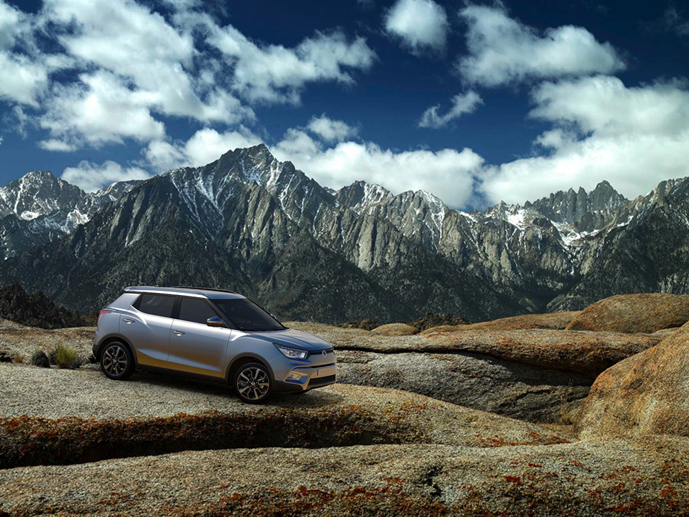 Silberner SUV auf felsigem Plateau vor imposanter Berglandschaft mit schneebedeckten Gipfeln unter blauem Himmel und Wolken.