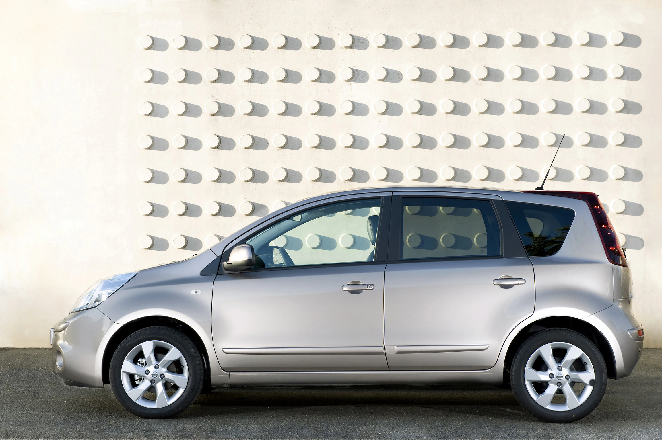 Silbernes Auto vor modern gestalteter Wand mit runden Aussparungen, seitlich betrachtet, auf einer Betonfläche geparkt.