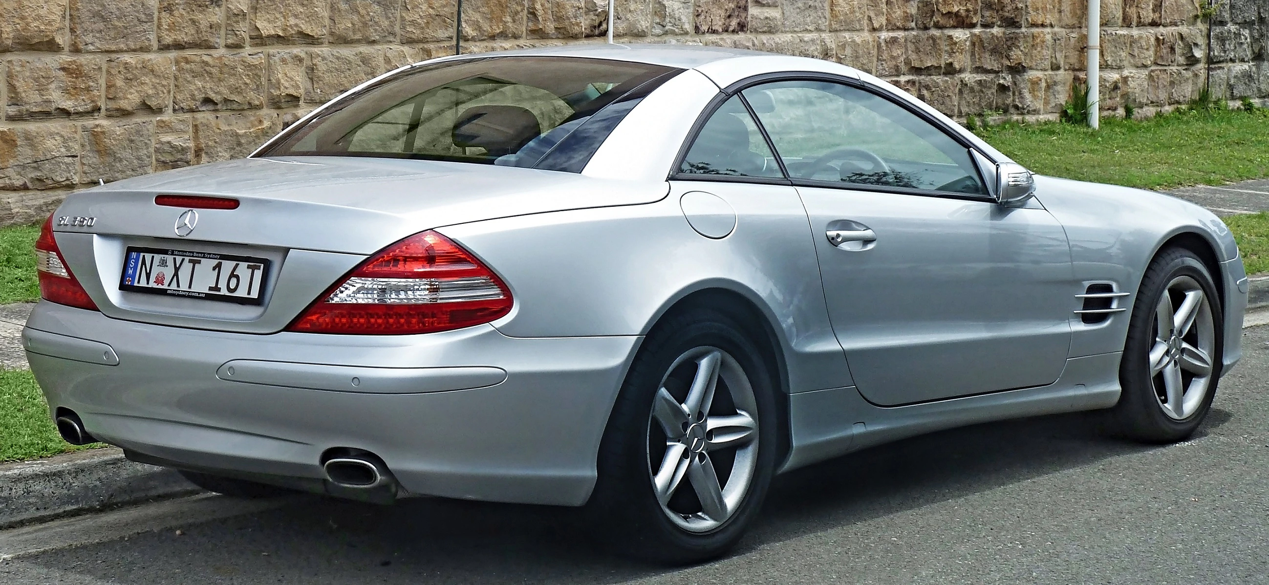 Silberner Mercedes-Benz SL 350 Cabrio, seitlich auf einer Straße geparkt, vor einer Ziegelmauer.