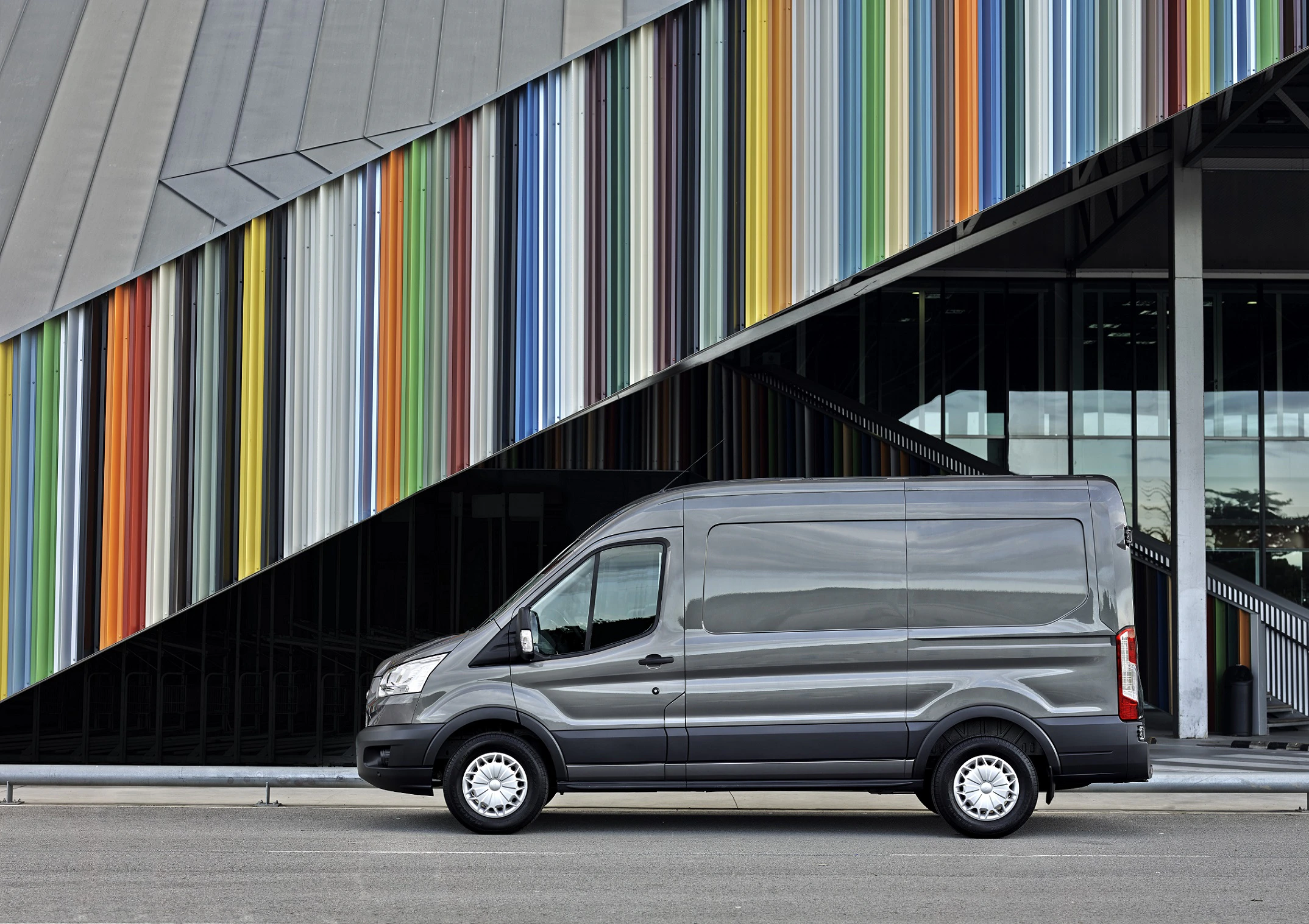 Grauer Transporter vor einem modernen Gebäude mit bunten vertikalen Linien und Glasfassade, die diagonale Strukturen erzeugt.