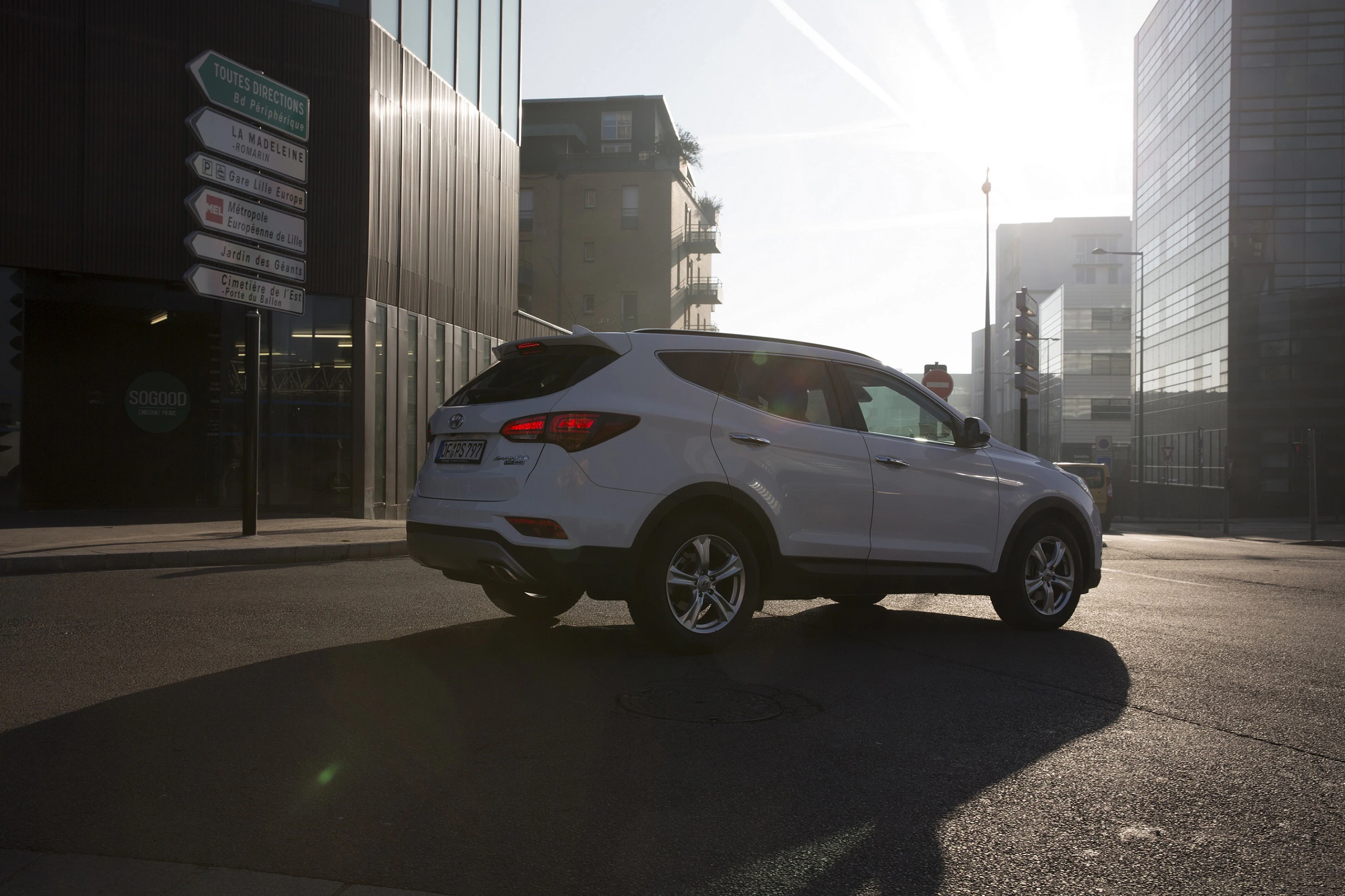 Ein weißes SUV fährt auf einer Stadtstraße bei Sonnenaufgang, umgeben von modernen Gebäuden und Straßenschildern.