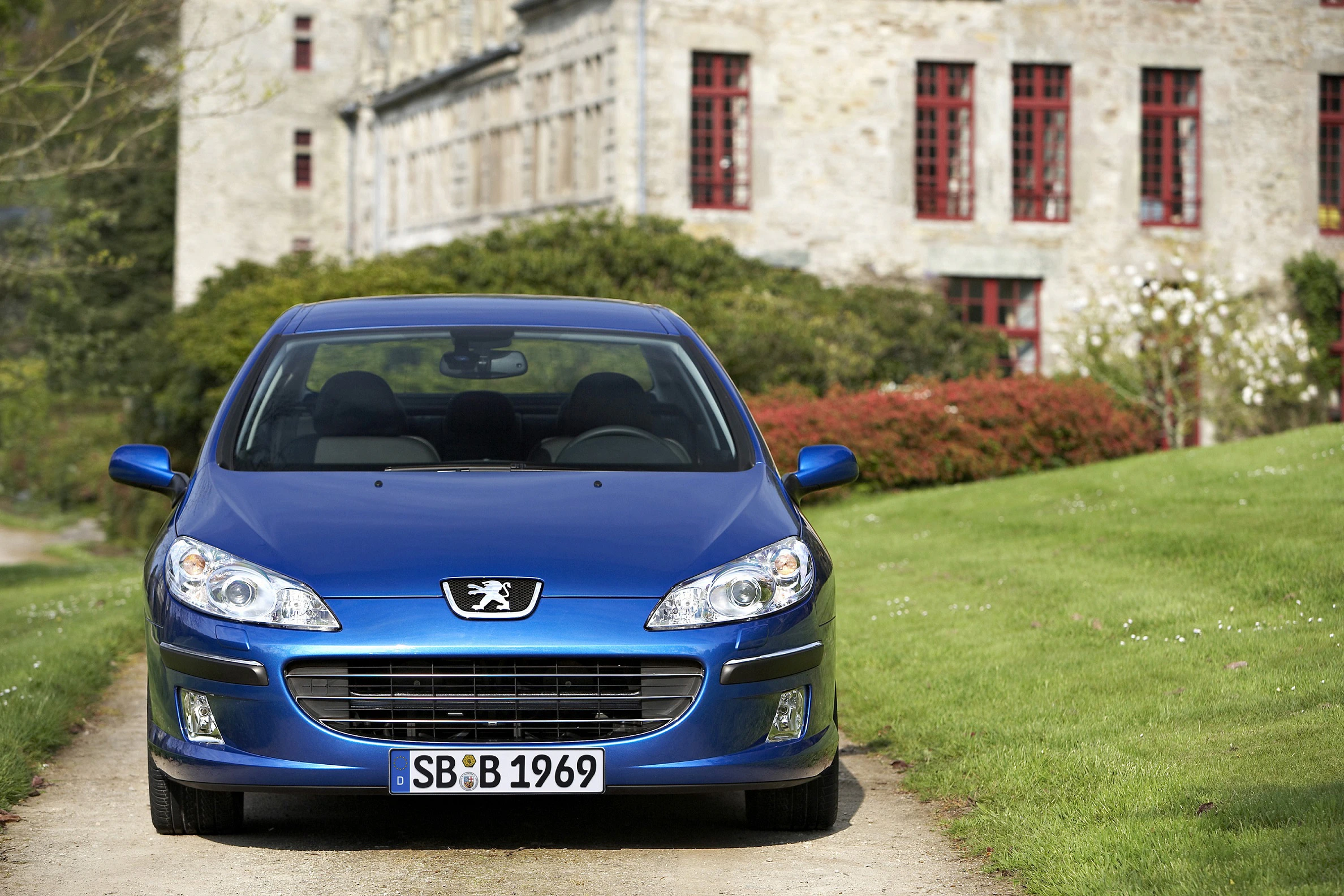 Blauer Peugeot auf einer Schotterstraße, im Hintergrund ein historisches Gebäude mit roten Fenstern.