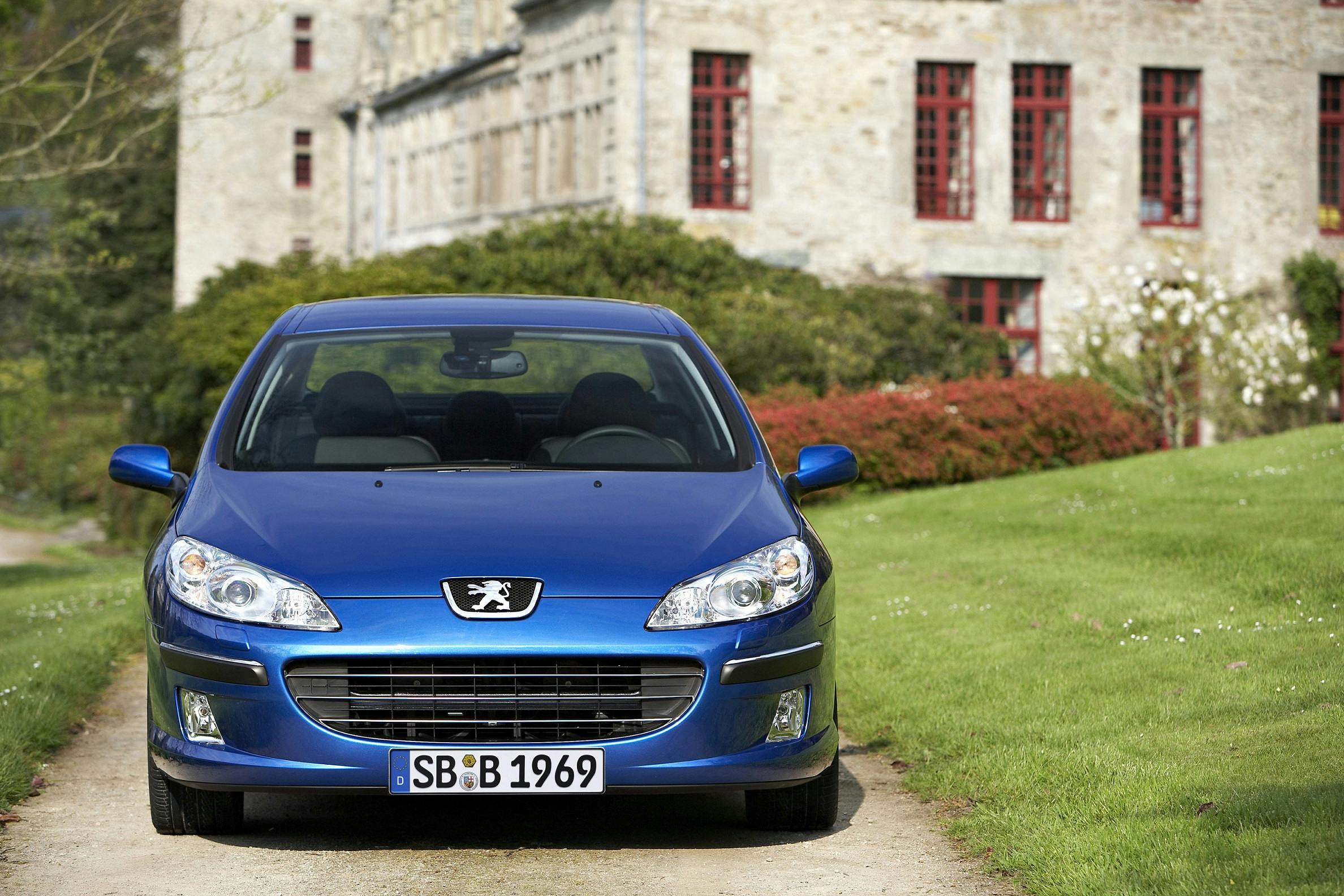 Blauer Peugeot auf einer Schotterstraße, im Hintergrund ein historisches Gebäude mit roten Fenstern.