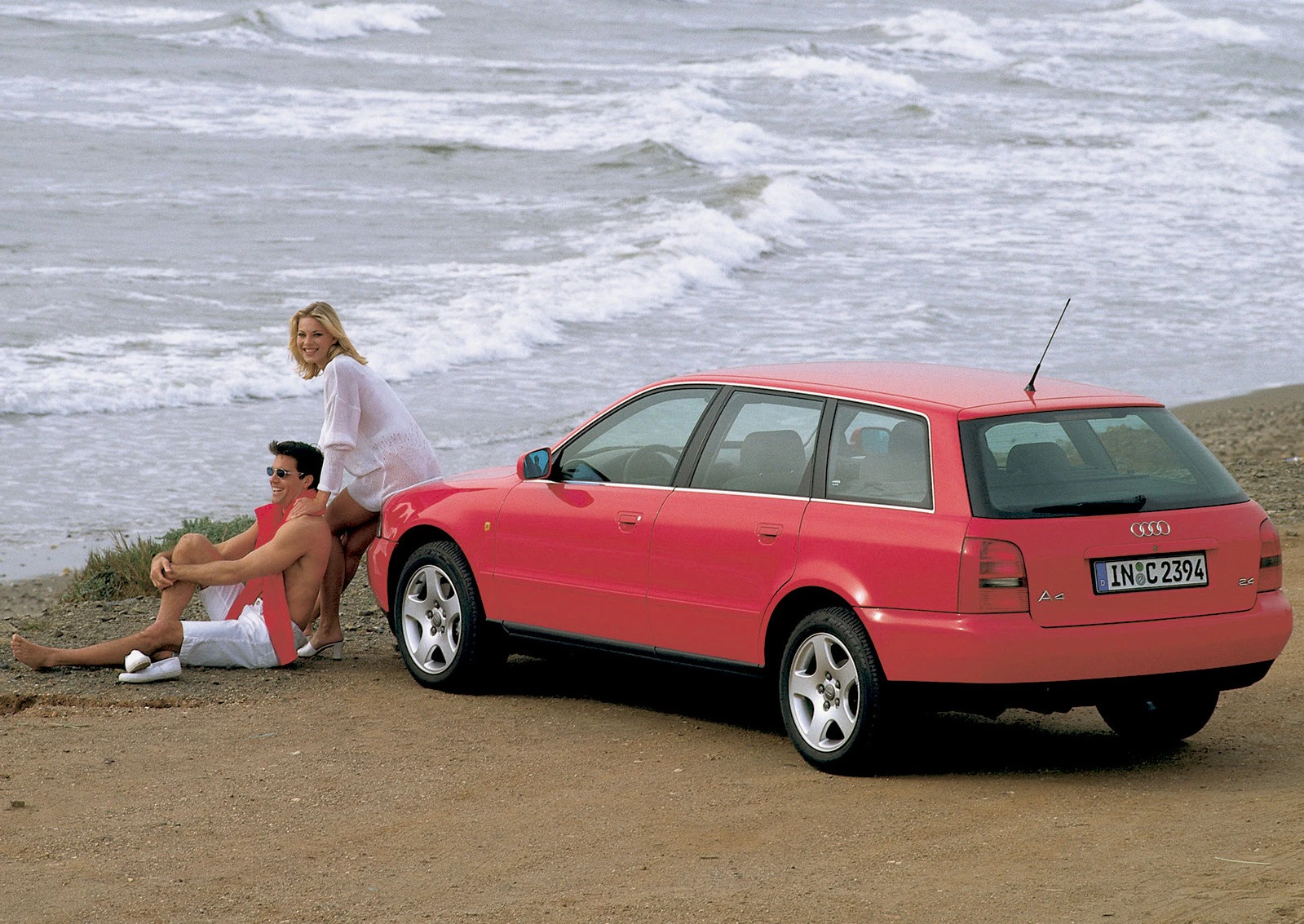 Roter Audi A6 Avant steht am Strand. Ein Mann und eine Frau sitzen lächelnd in Badebekleidung in der Nähe des Autos, Meer im Hintergrund.