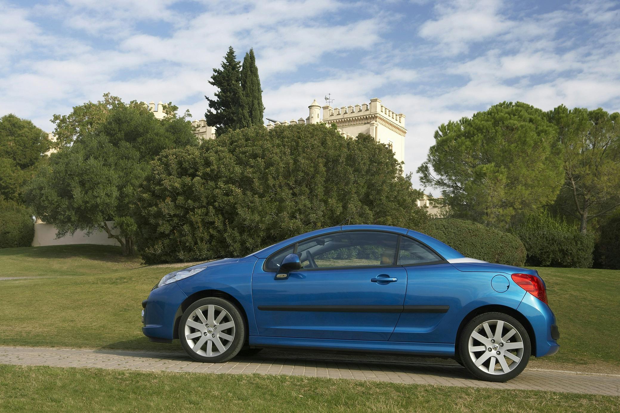 Blaues Cabrio geparkt auf einer Rasenfläche vor einem großen, historischen Gebäude mit grünem Garten und bewölktem Himmel im Hintergrund.