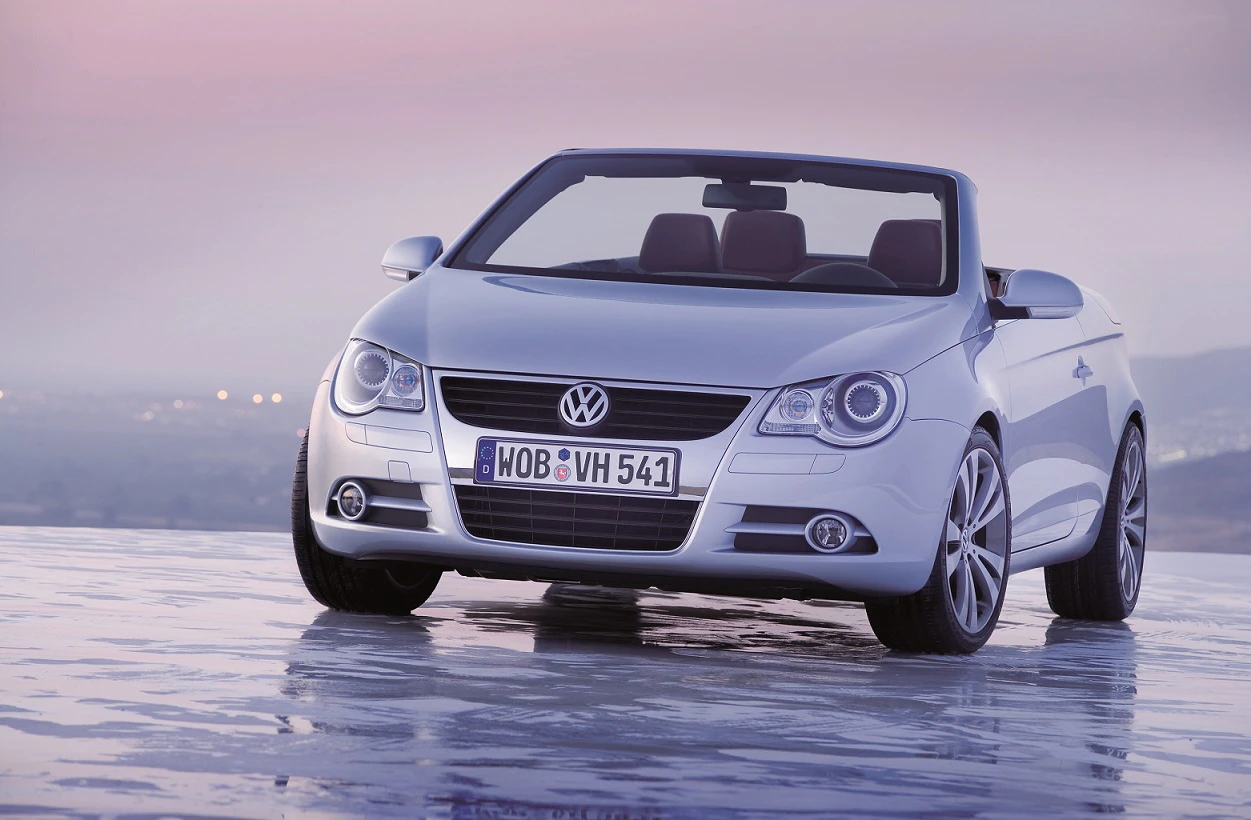 Silberner Volkswagen-Cabrio parkt auf nasser Oberfläche, Frontansicht. Hintergrund verschwommen, Himmel rosa-grau.