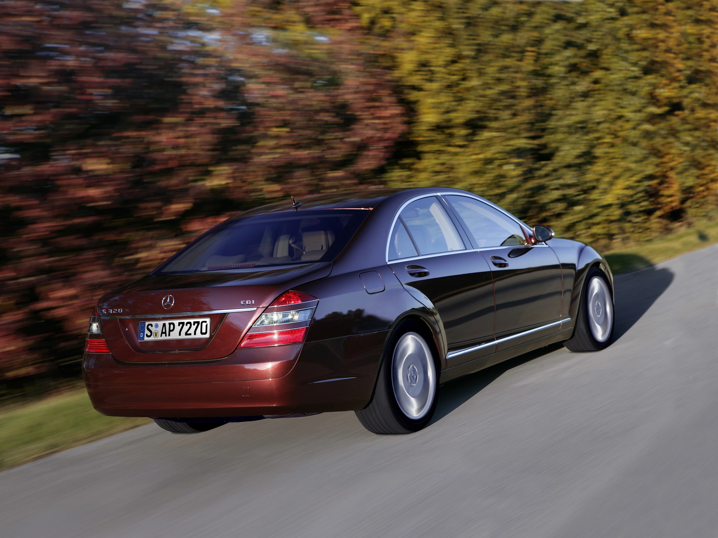 Brauner Mercedes-Benz S-Klasse fährt auf einer Straße, umgeben von herbstlichen Bäumen im Hintergrund.