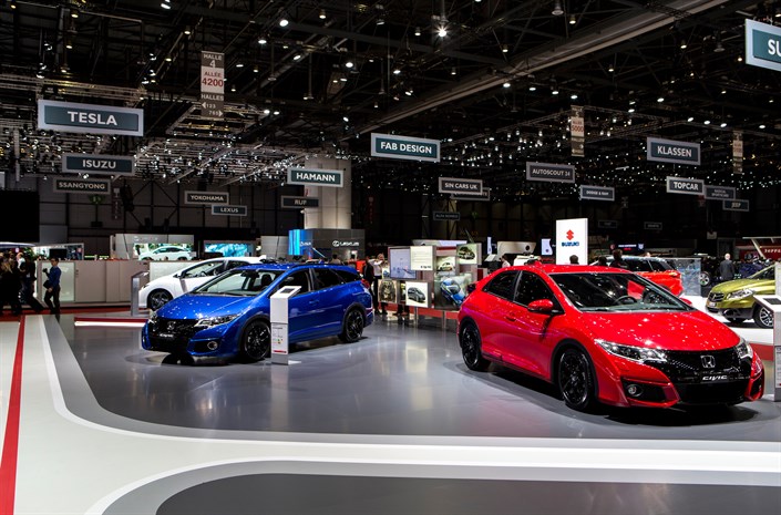 Autoshow mit leuchtend roten und blauen Autos auf glänzendem Boden, umgeben von verschiedenen Markenständen in einer großen Messehalle.