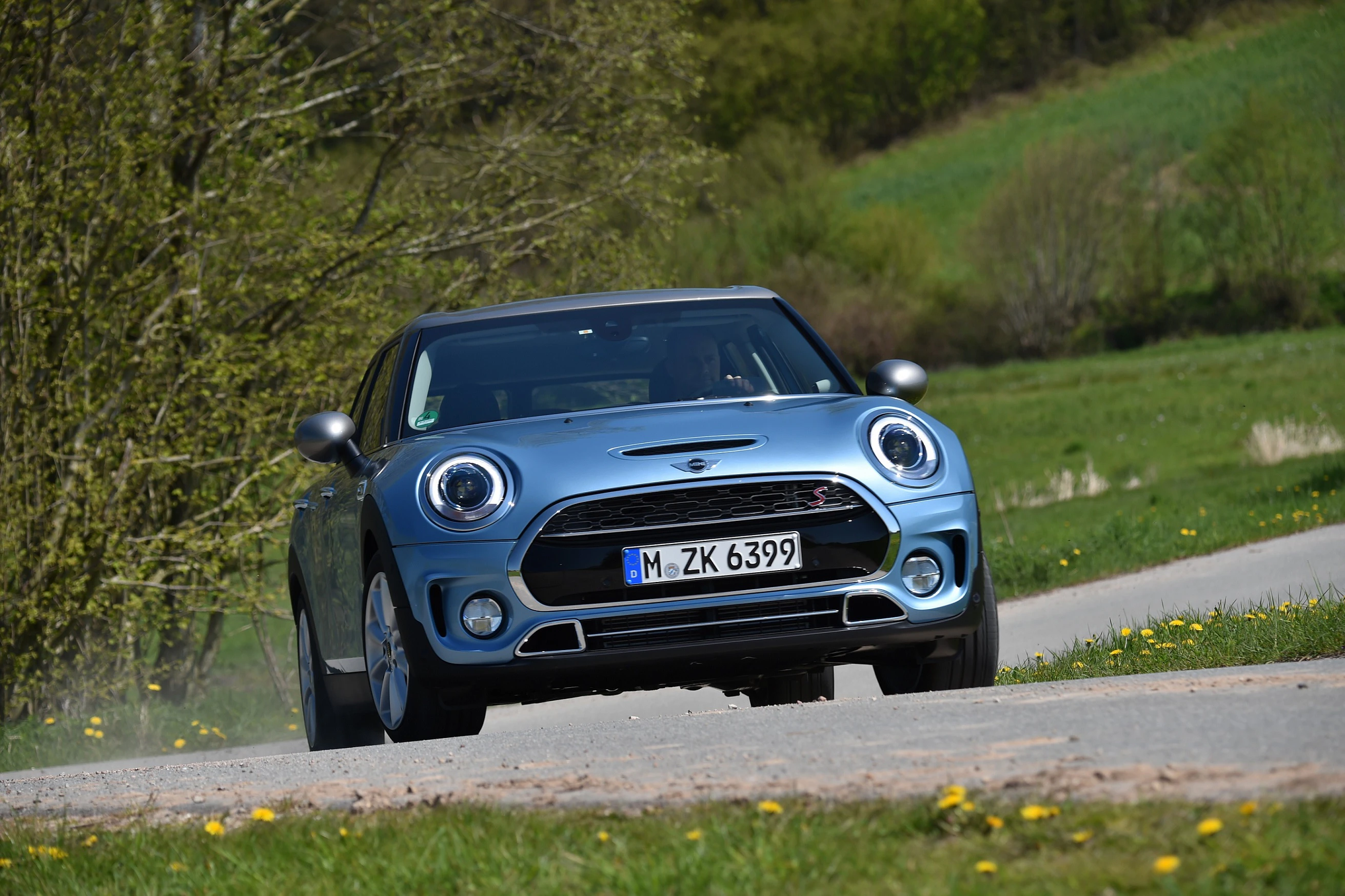 Blauer Mini Cooper fährt auf ländlicher Straße. Umgeben von grünen Feldern und Bäumen, sonniger Tag.