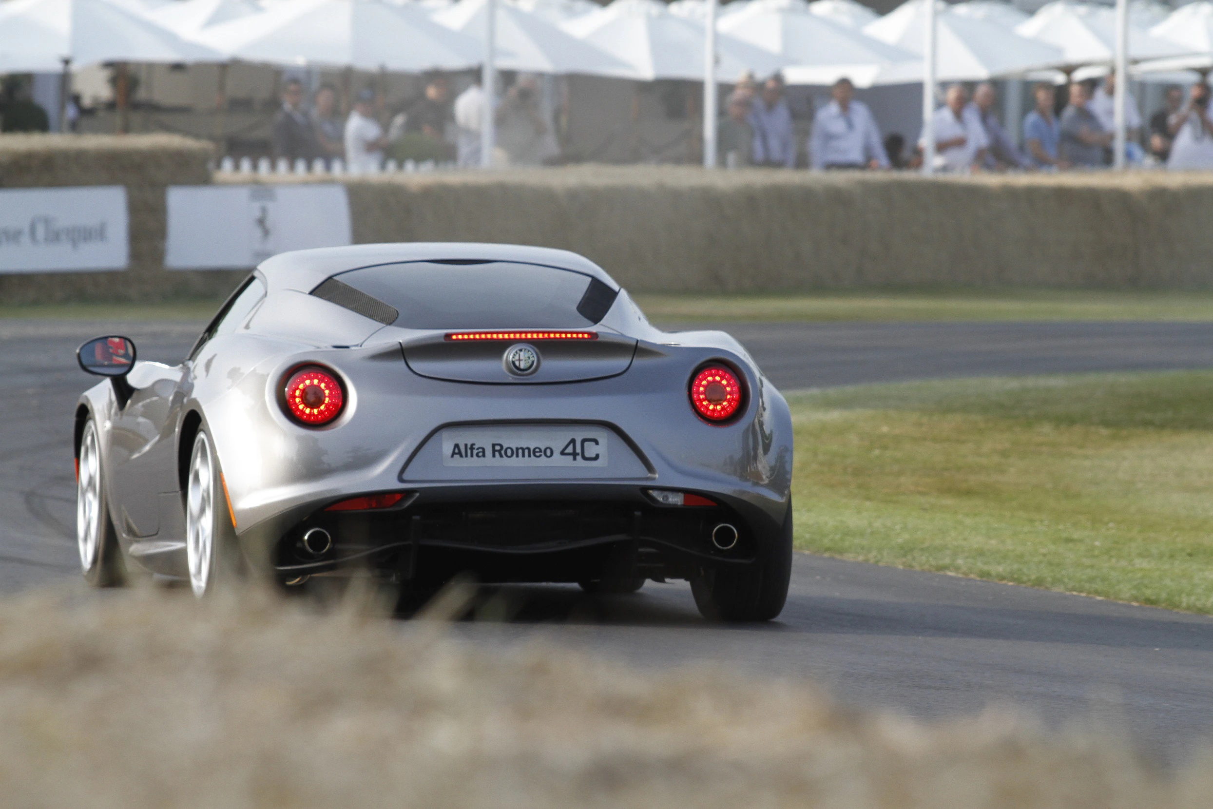 Silberner Alfa Romeo 4C fährt auf einer Rennstrecke, Zuschauer im Hintergrund.