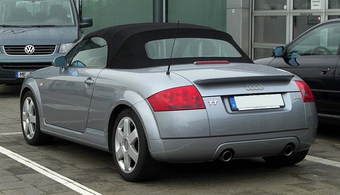 Silberner Audi TT Cabrio auf einem Parkplatz, Heckansicht. Parkplatz mit mehreren Autos im Hintergrund.