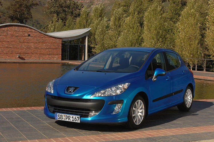Blauer Peugeot 308 steht auf gepflasterter Fläche vor einem modernen Gebäude mit Bäumen im Hintergrund.