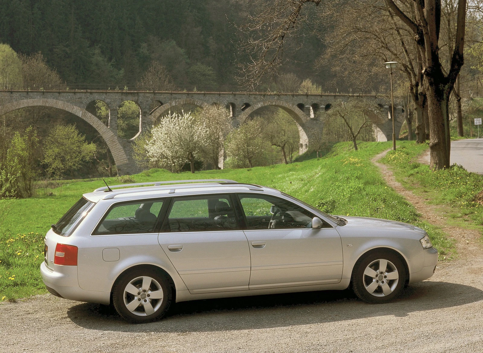 Silberner Kombi auf einem Parkplatz vor einer steinernen Eisenbahnbrücke und grüner Landschaft mit Bäumen im Hintergrund.