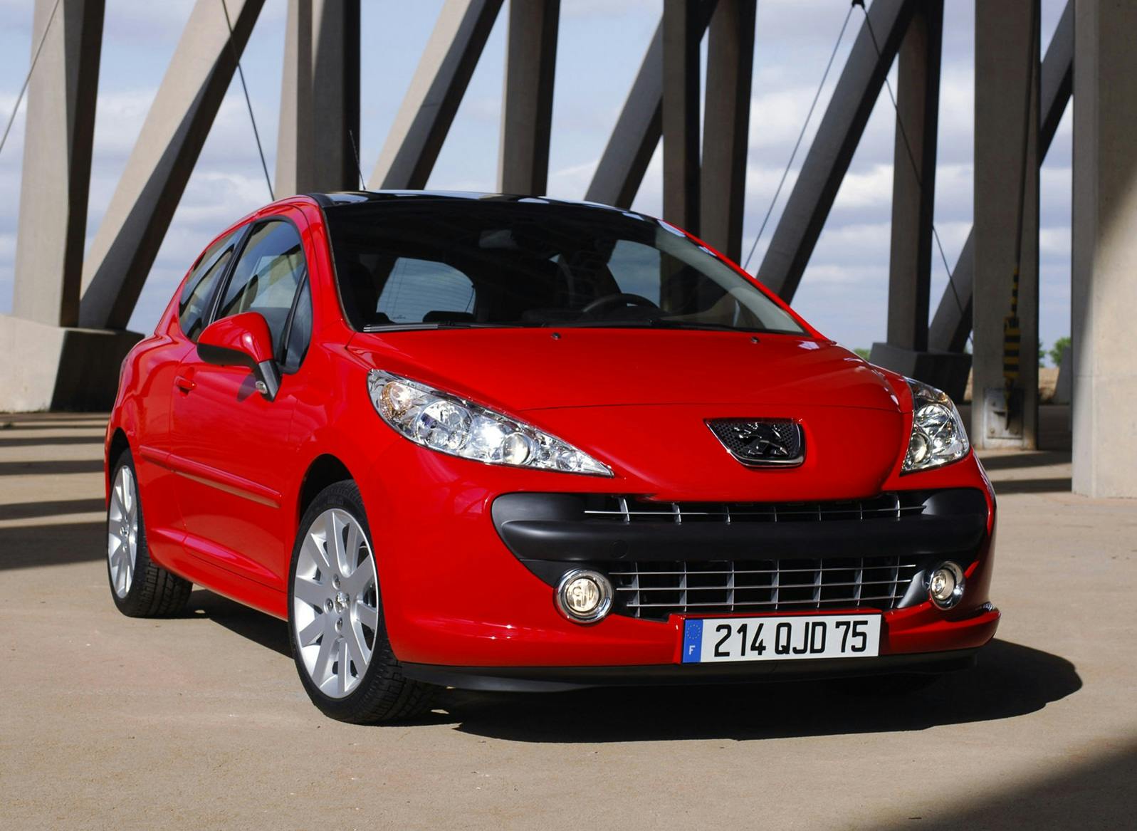 Roter Peugeot 207 steht auf einem Parkplatz vor einer modernen Stahlstruktur, bei Tageslicht.