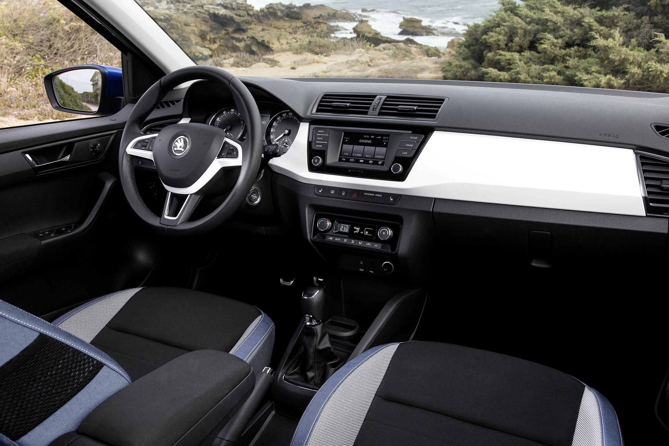 Innenraum eines Autos mit modernem Lenkrad, Armaturenbrett und Infotainmentsystem, im Hintergrund Küstenlandschaft sichtbar.