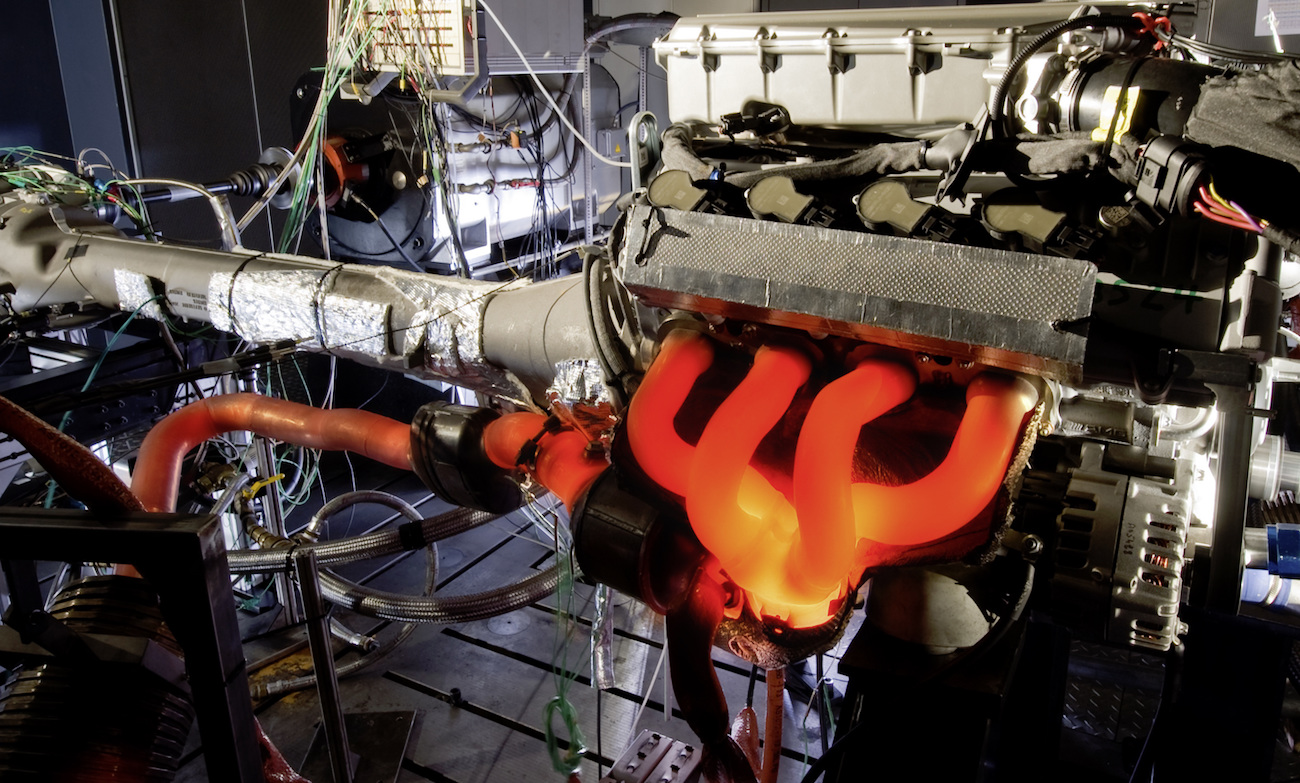 Glühender Autoauspuffkrümmer in einer Werkstatt, umgeben von Kabeln und Maschinen, zeigt intensiven Motorbetrieb.