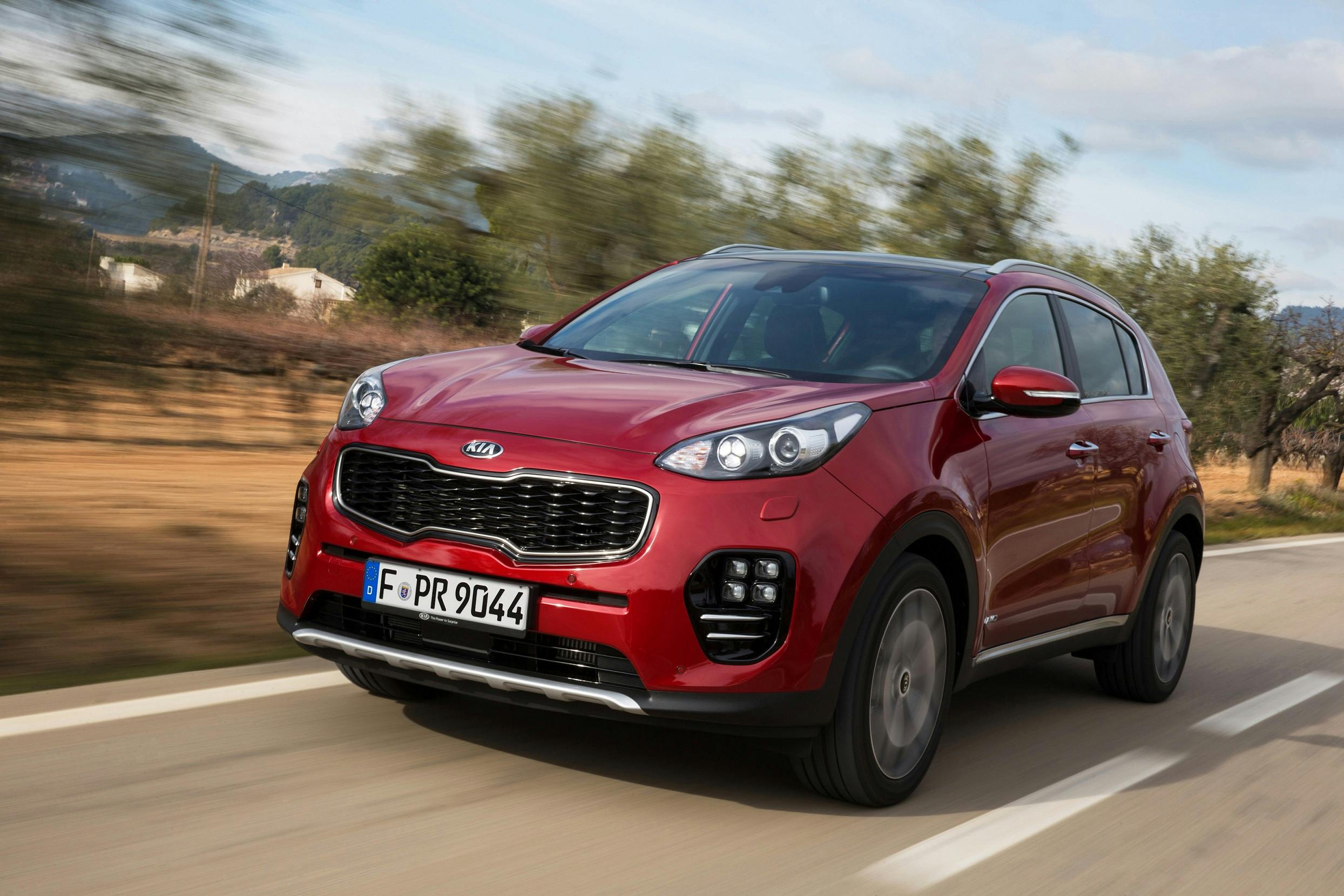 Roter Kia Sportage SUV fährt auf einer Landstraße, umgeben von Natur und bewölktem Himmel.